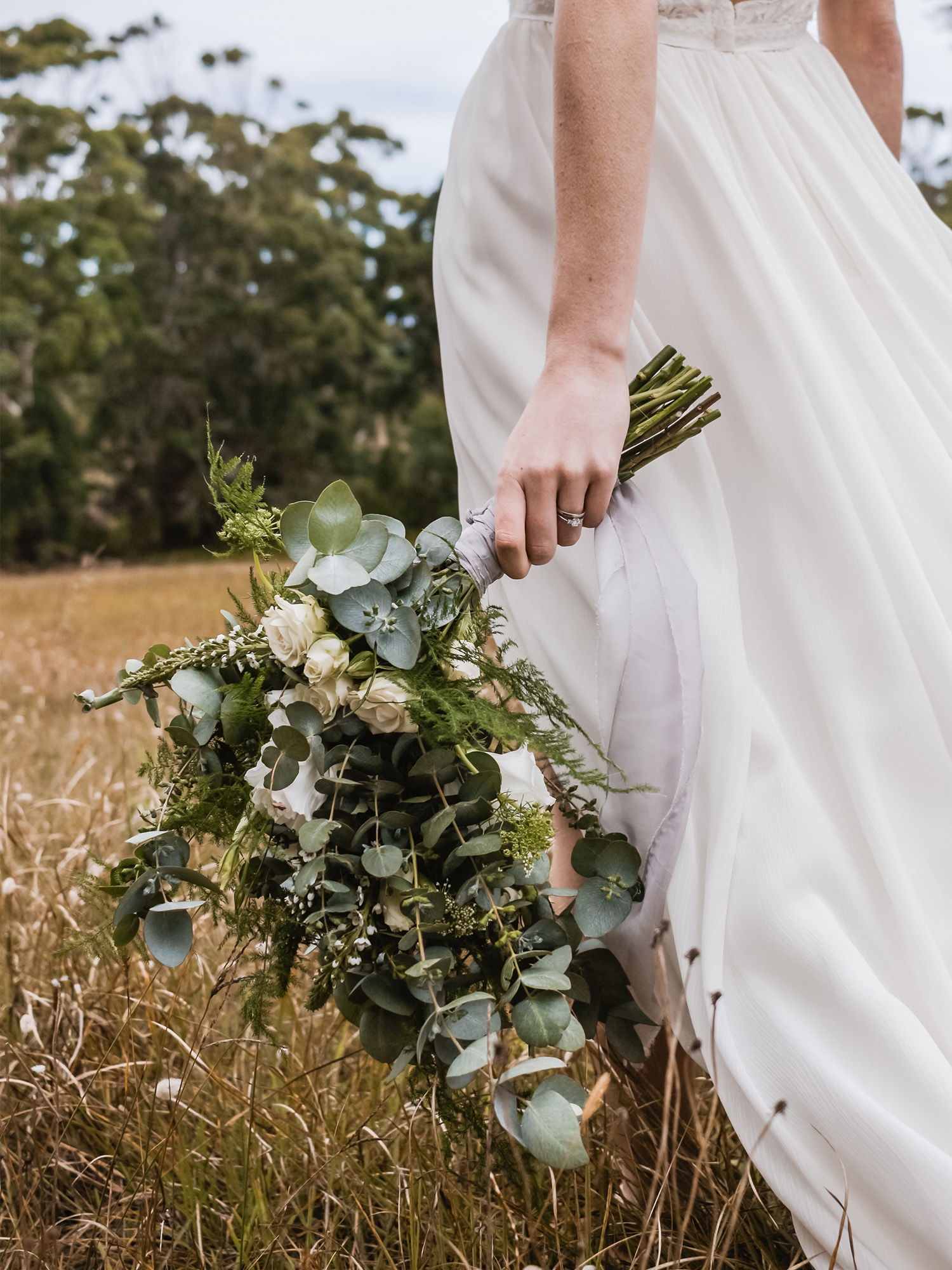 Nahaufnahme der Hand einer Braut, die einen rustikalen Strauß mit Eukalyptusblättern und weißen Blumen festhält, während sie in einer ländlichen Umgebung steht.