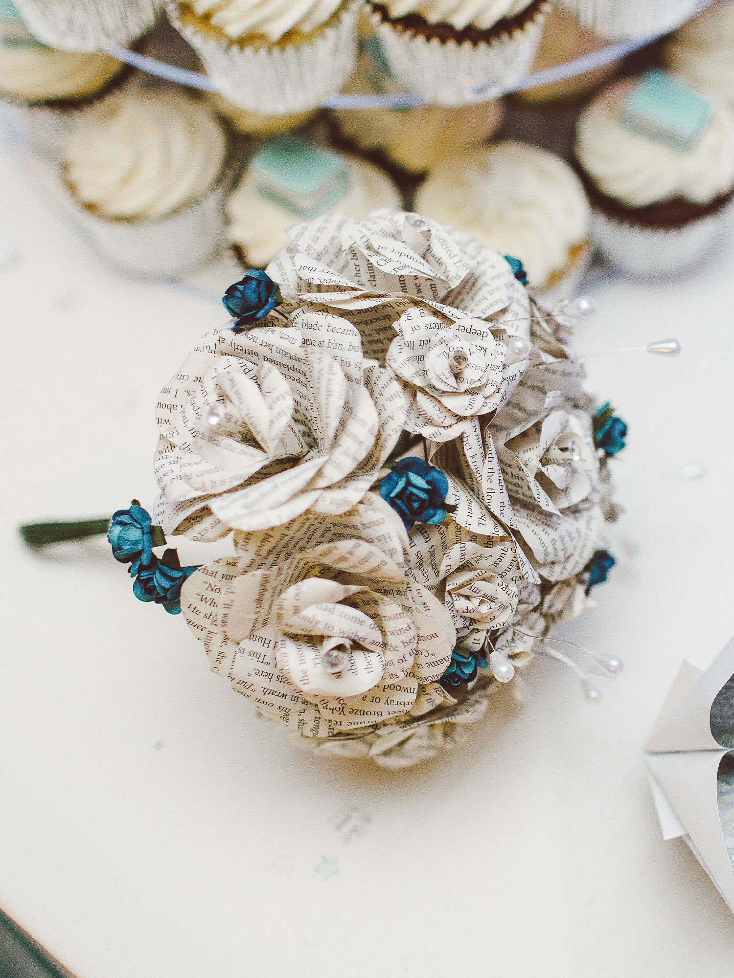 Handgefertigter Brautstrauß aus Buchseiten-Rosen mit blauen Blüten und Perlendekorationen, arrangiert neben Cupcakes als festliche Tischdeko.