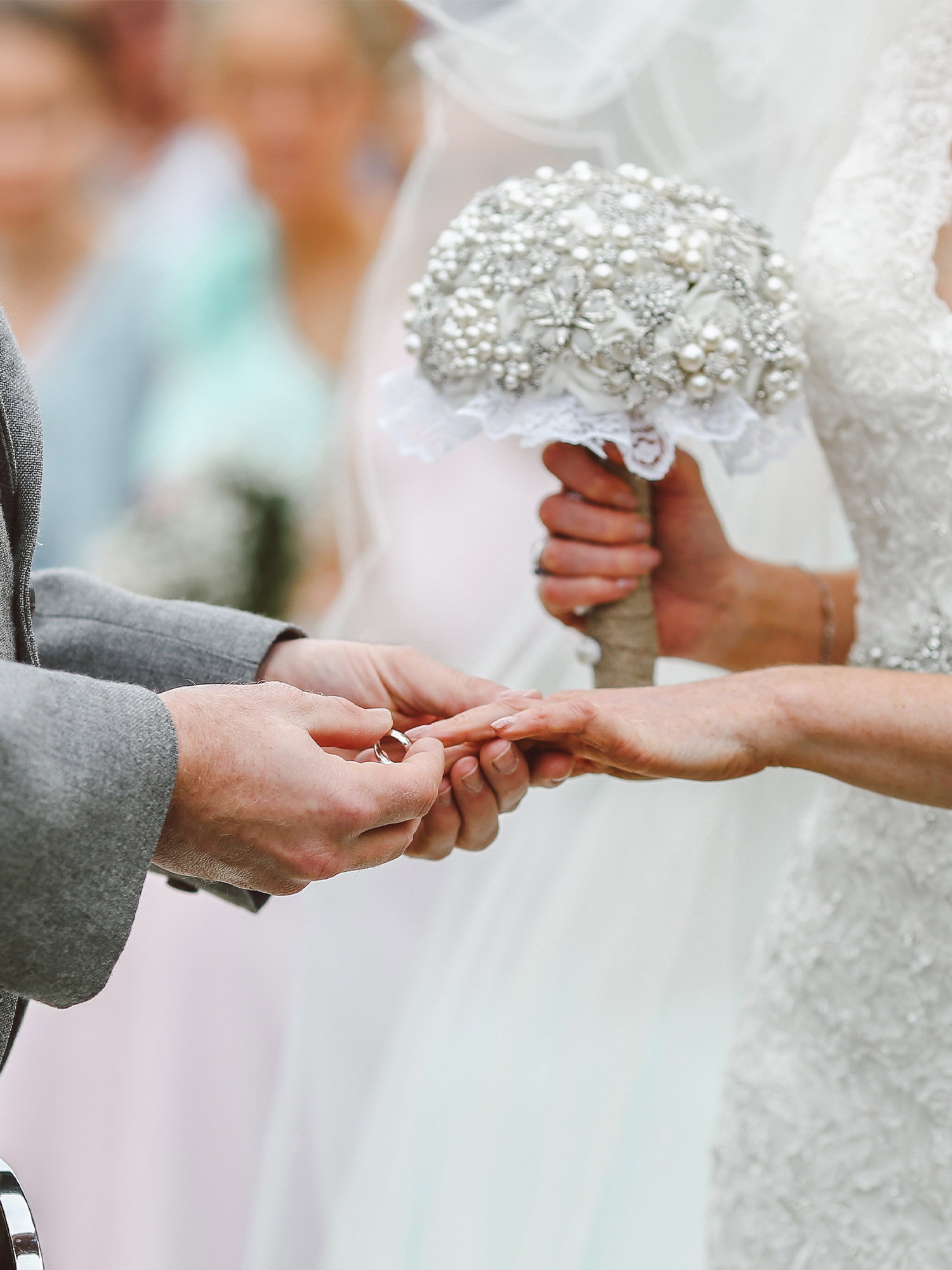 Nahaufnahme der Hände des Brautpaares beim Ringtausch, die Braut trägt ein elegantes Kleid und hält einen glitzernden Strassstrauß.