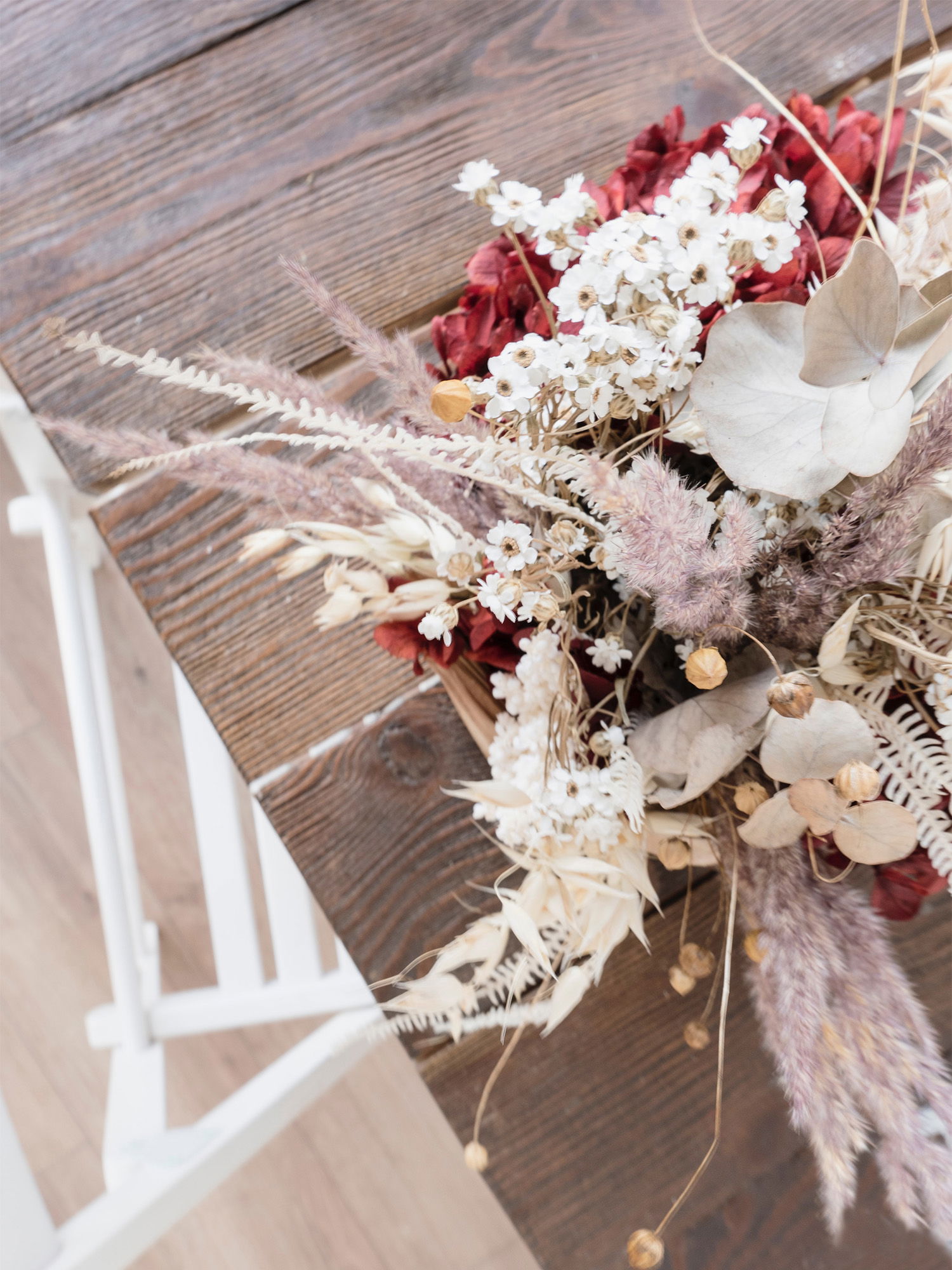 Ein eleganter Brautstrauß aus getrockneten Blumen in Weiß- und Rottönen, präsentiert auf einer hölzernen Tischplatte.