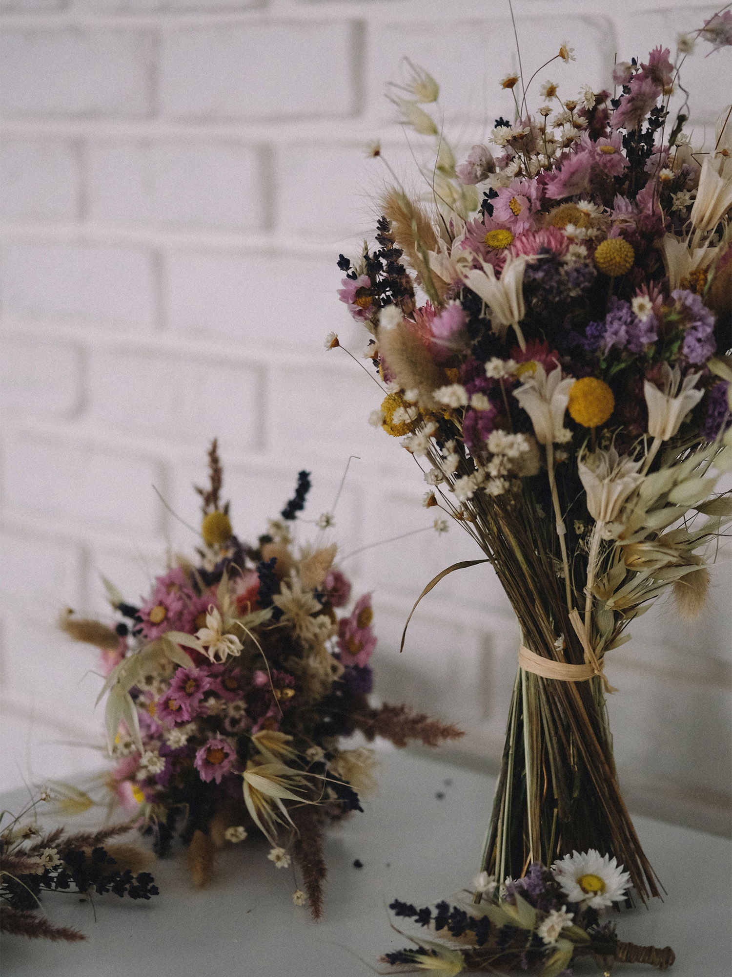 Zwei dekorative Sträuße aus Trockenblumen mit lila, gelben und weißen Blüten, stilvoll vor einer hellen Backsteinwand platziert.