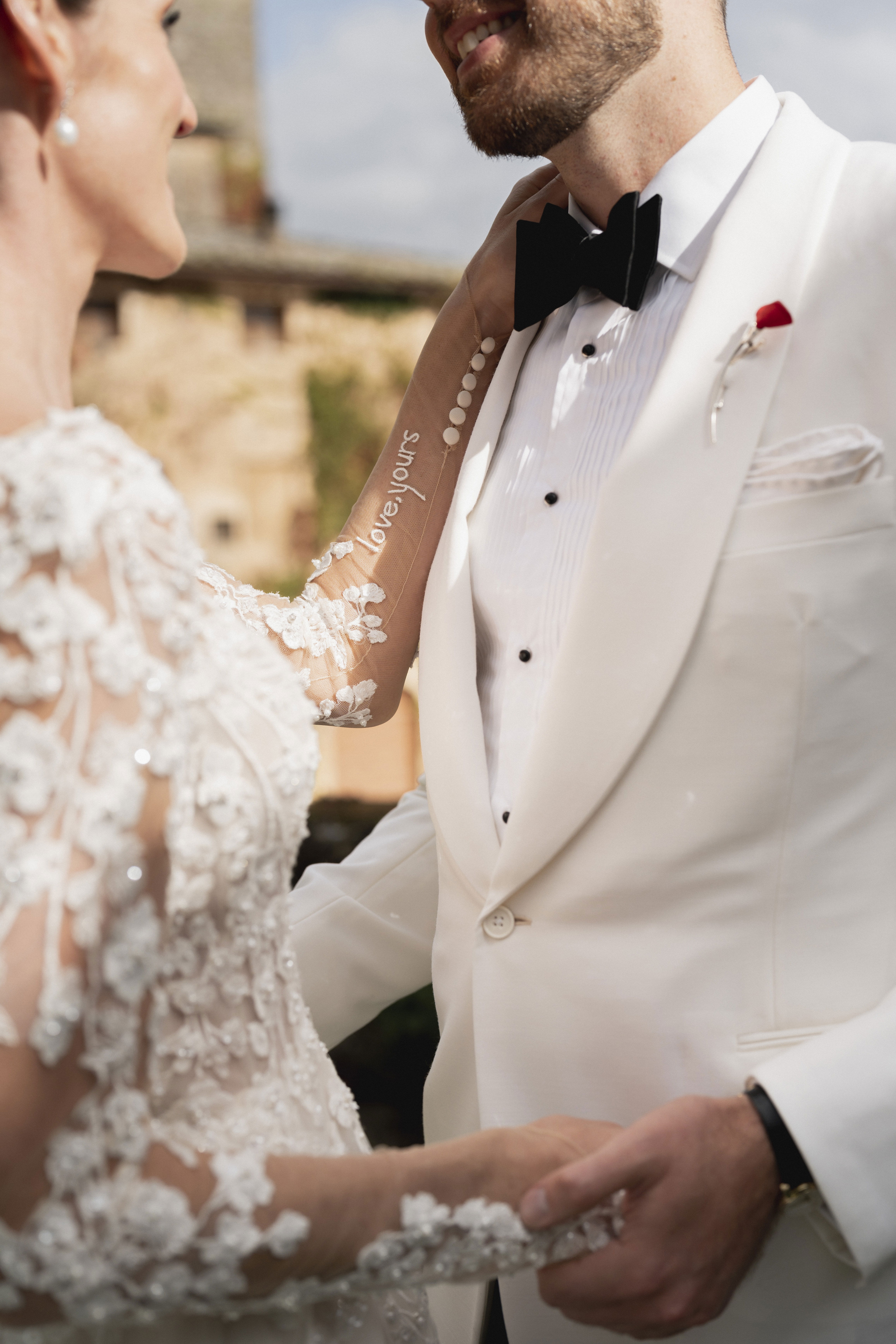 Detailansicht des Brautpaares, wie sie sich zärtlich an den Händen halten. Der Bräutigam trägt einen eleganten weißen Anzug, während die Braut ein aufwendig besticktes Kleid mit floralen Spitzen und einem besonderen Schriftzug auf dem Ärmel trägt.