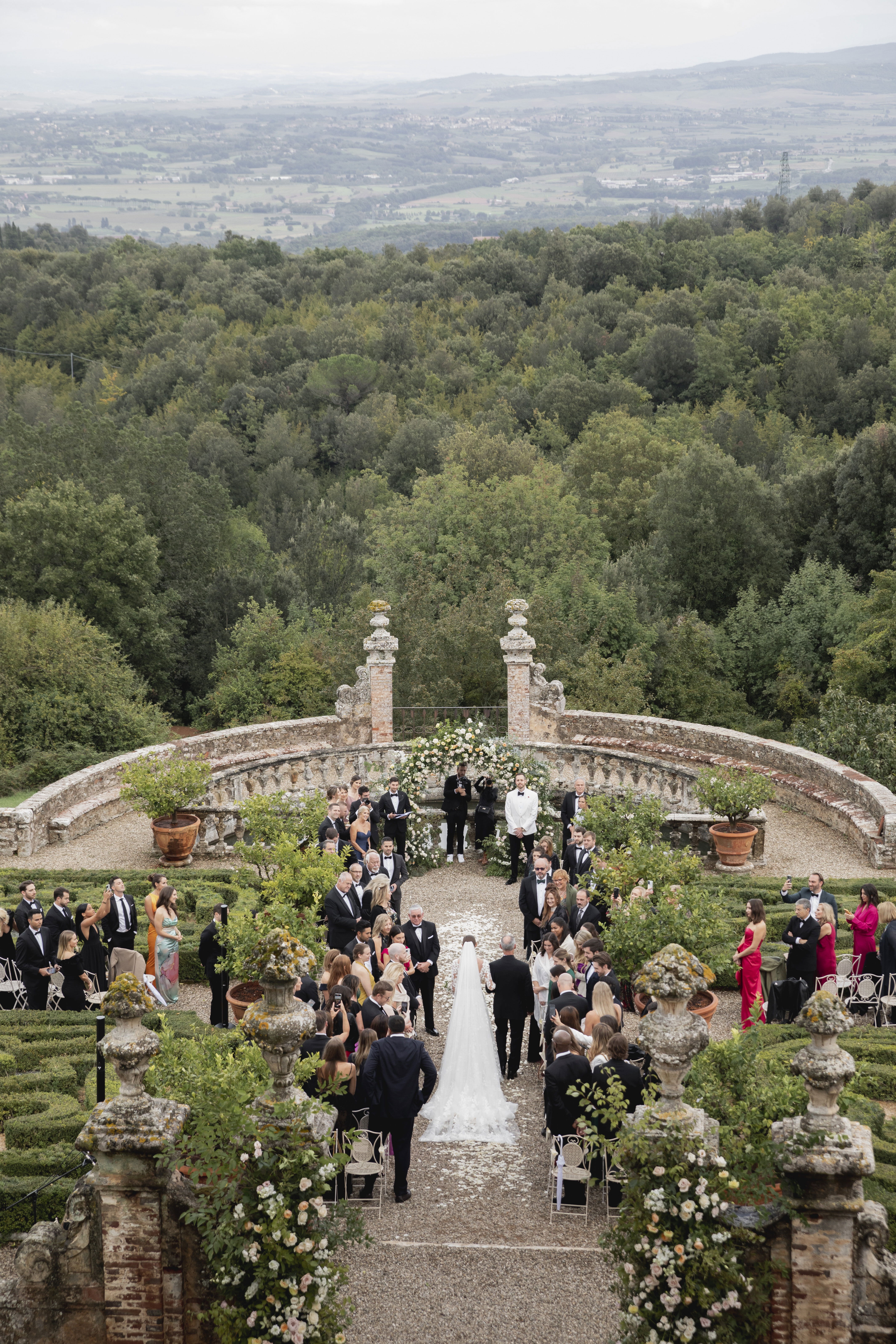 Romantische Hochzeitszeremonie unter freiem Himmel, mit einem von Blumen verzierten Altar und einer großen Anzahl von Gästen. Die Braut, in einem langen Schleier, geht den Gang entlang, während die Zeremonie inmitten einer malerischen Landschaft stattfindet.