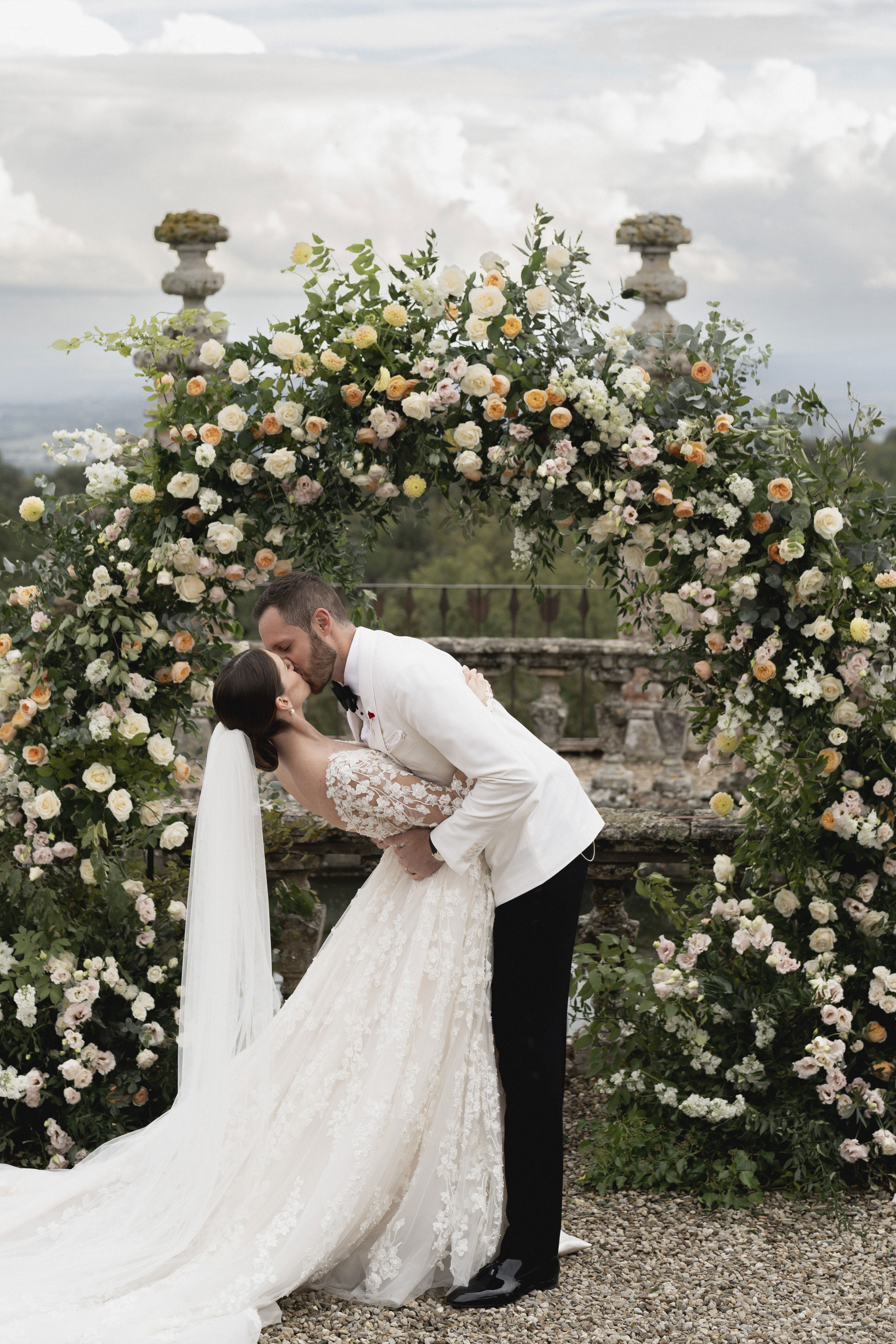 Ein Hochzeitspaar teilt einen Kuss unter einem reich verzierten Blumenbogen, der mit weißen, gelben und rosa Blüten geschmückt ist. Die Braut trägt ein aufwendig verziertes Spitzenkleid mit Schleier, während der Bräutigam einen weißen Smoking trägt.