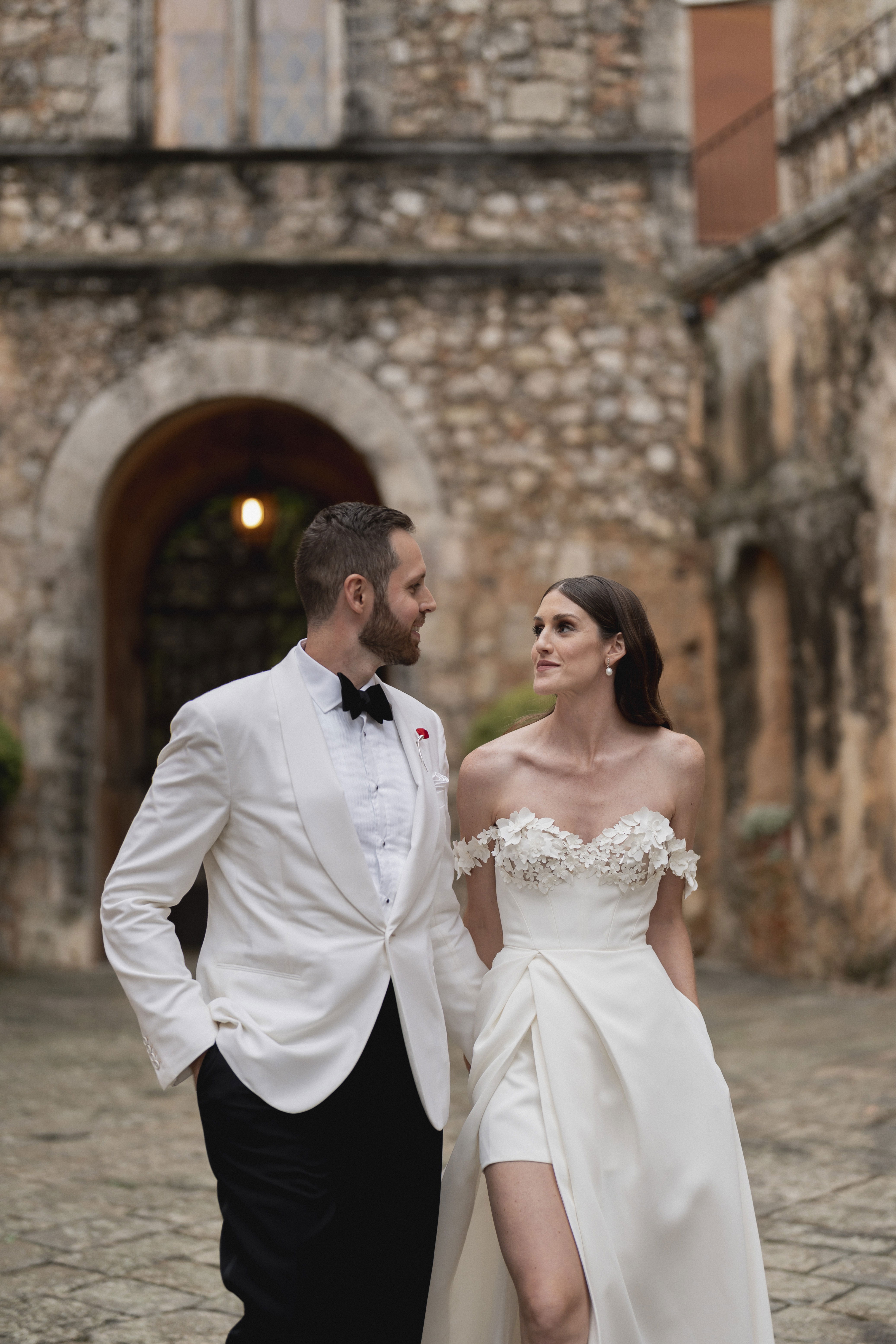 Romantischer Spaziergang eines Hochzeitspaares in einem alten Steinbau. Die Braut trägt ein modernes, schulterfreies Kleid mit Blumenapplikationen und der Bräutigam einen eleganten weißen Smoking.