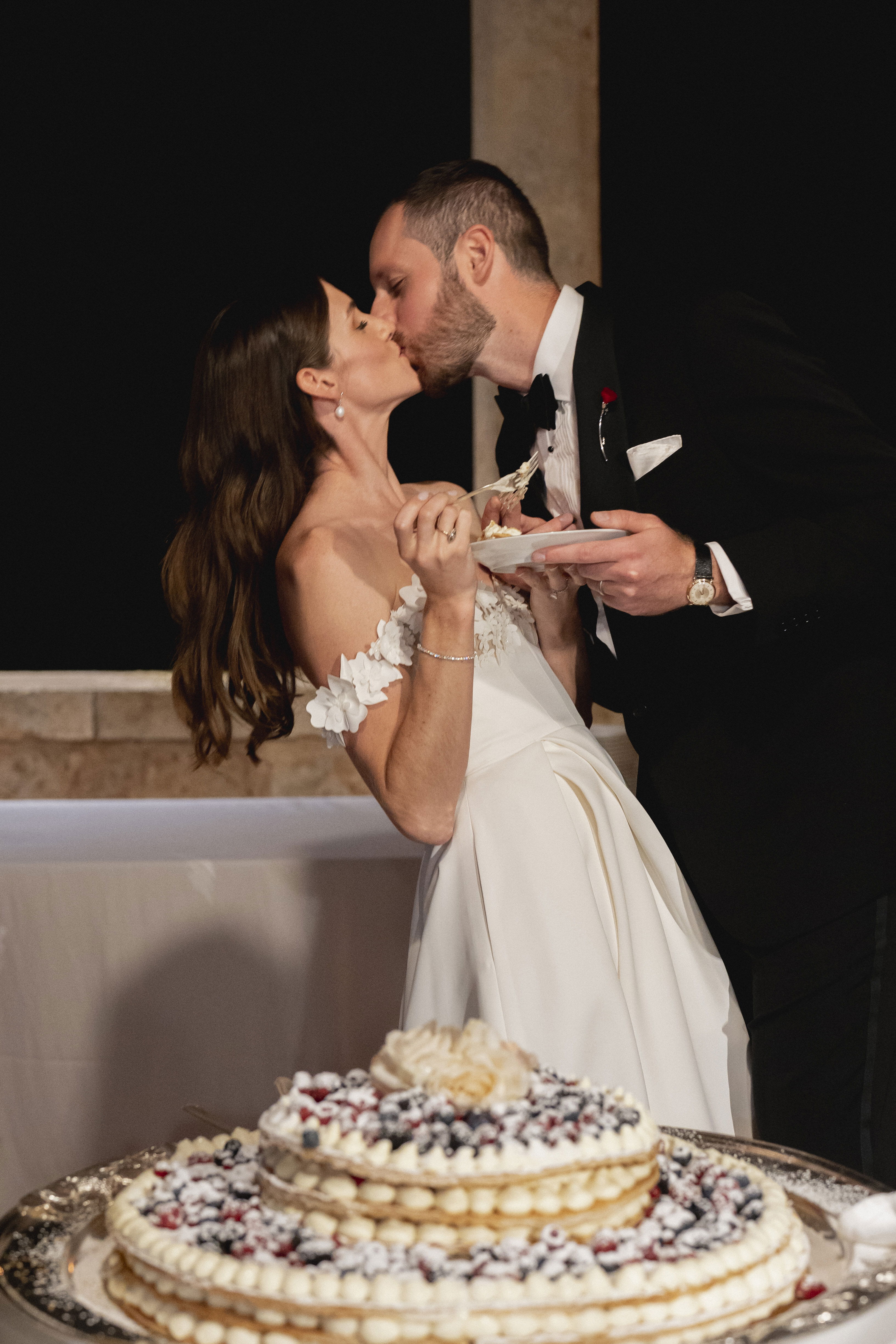 Das Brautpaar küsst sich romantisch, während sie gemeinsam ein Stück ihrer Hochzeitstorte genießen. Die Braut in einem schulterfreien Kleid und der Bräutigam im Smoking halten jeweils Teller mit Kuchen in den Händen, im Vordergrund ist die dekorierte Torte zu sehen.