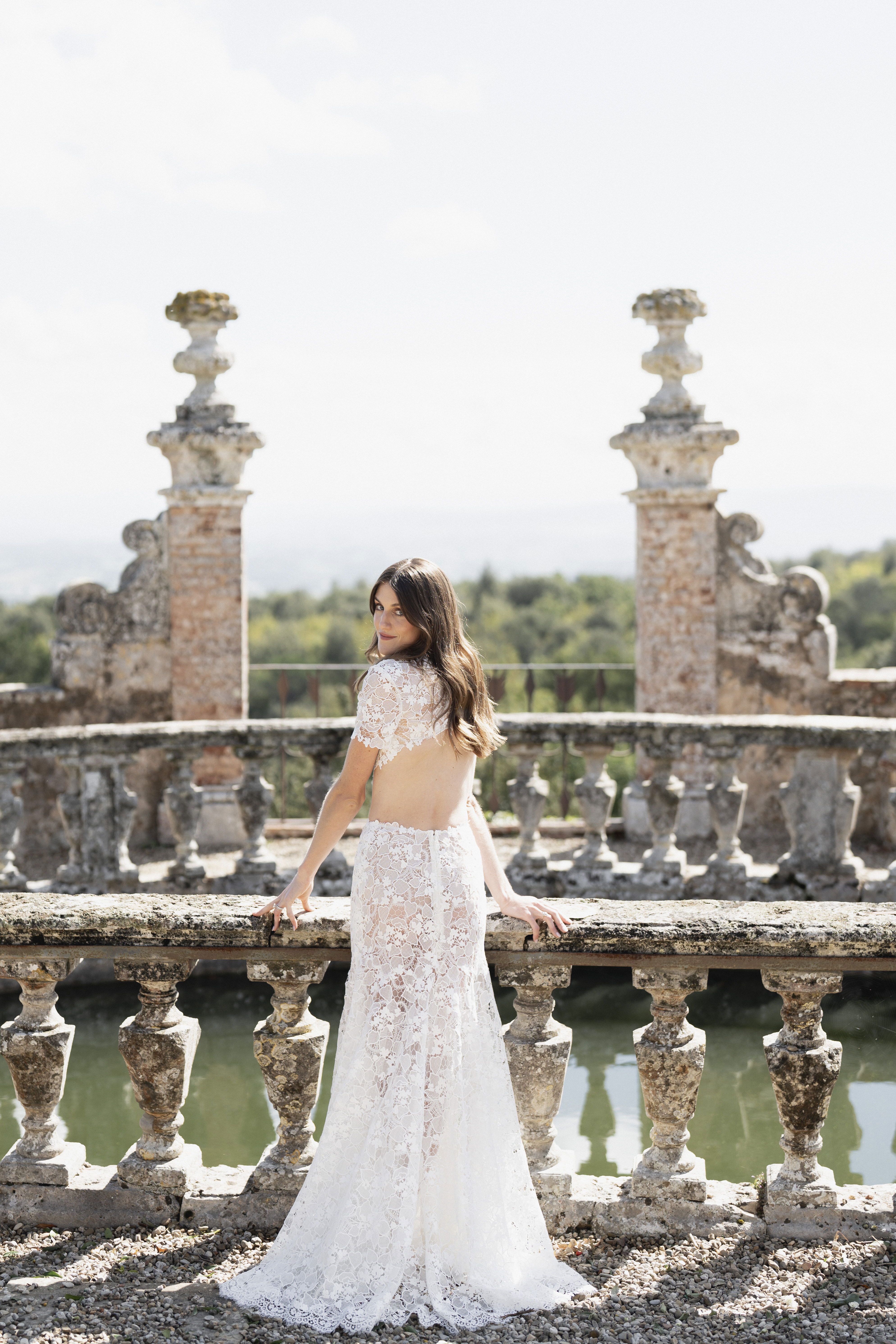 Die Braut steht in einem eleganten, rückenfreien Spitzenkleid auf einer historischen Terrasse und blickt über die Schulter in die Kamera. Die filigrane Spitze des Kleides betont die romantische Szenerie vor einer malerischen Kulisse.