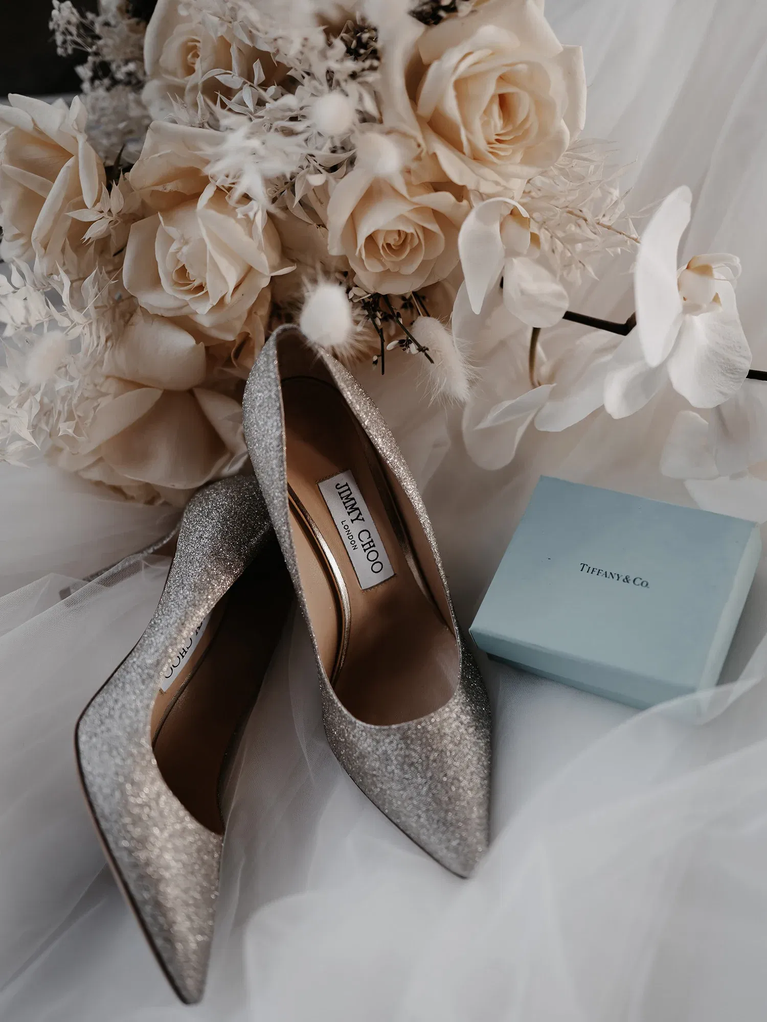 Brautschuhe in funkelndem Silber von Jimmy Choo, arrangiert mit einem Strauß aus elfenbeinfarbenen Rosen und einer ikonischen blauen Tiffany & Co. Schachtel.