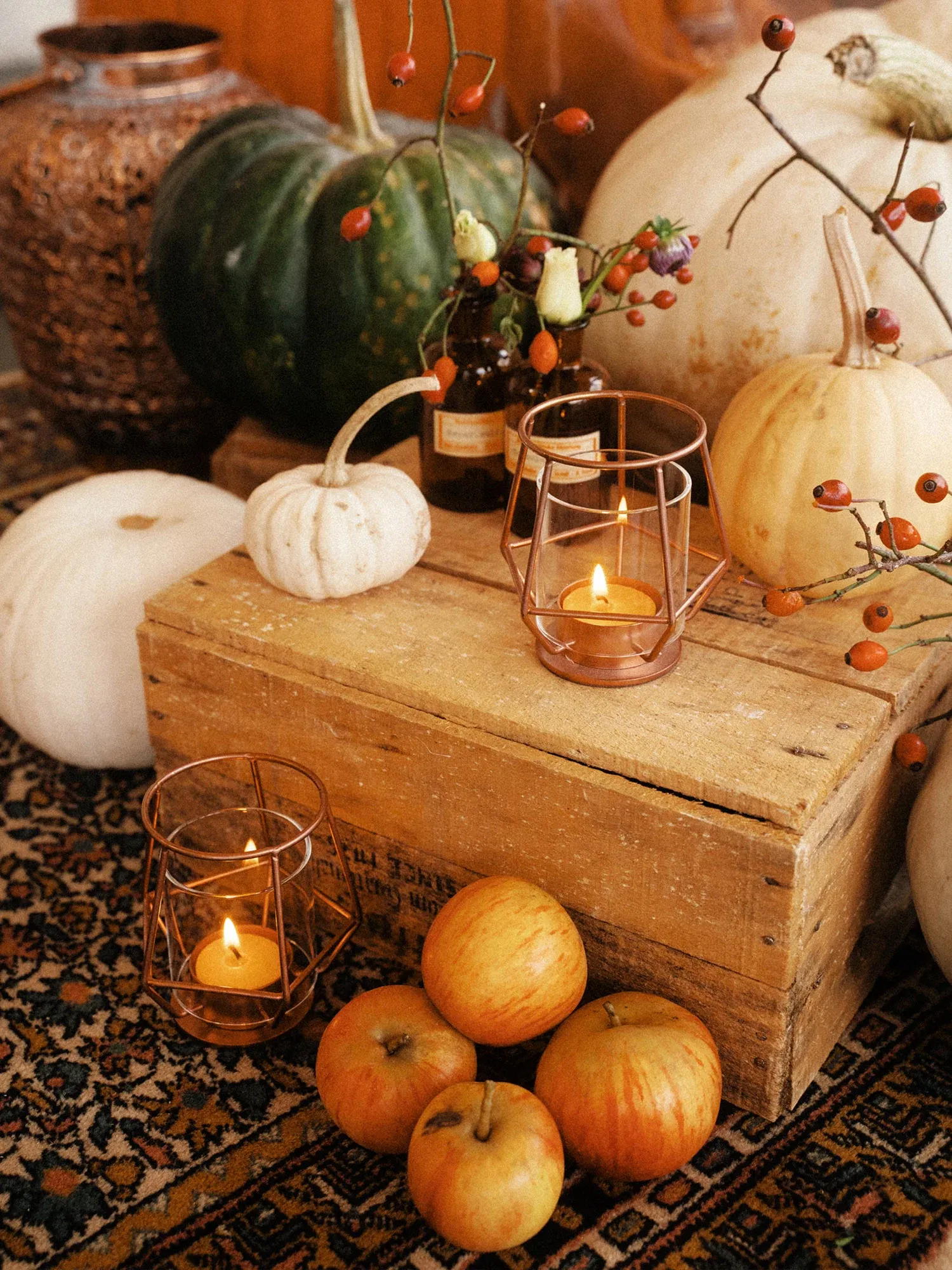 Herbstliche Dekoration mit Kürbissen, Äpfeln, Kerzen in kupferfarbenen Laternen und Zweigen auf einer rustikalen Holzkiste.