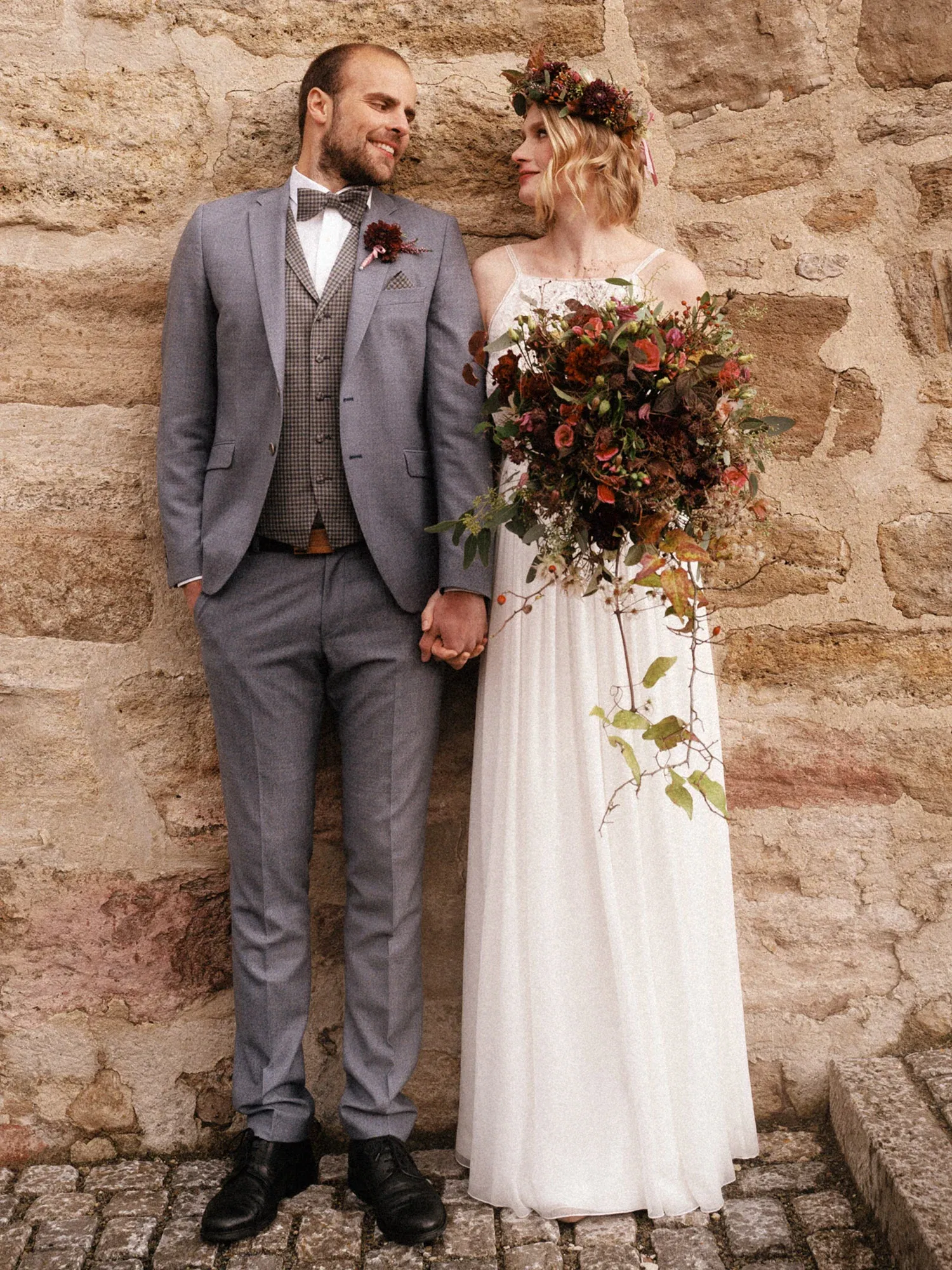 Ein Brautpaar lehnt an einer Sandsteinwand. Die Braut trägt ein Spitzenkleid mit Blumenkranz und Brautstrauß, der Bräutigam einen grauen Anzug.