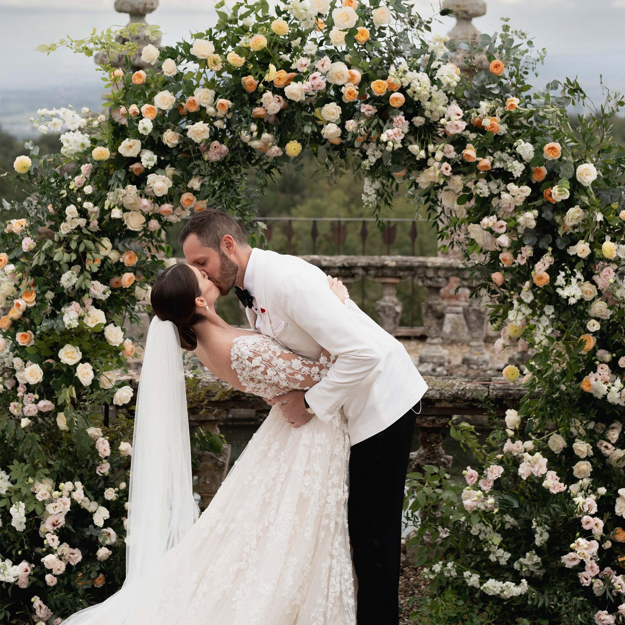 Braut und Bräutigam küssen sich vor einem wunderschönen, blumengeschmückten Traubogen im Freien. Die Braut ist in einem eleganten Spitzenkleid mit langem Schleier gekleidet, während der Bräutigam einen eleganten weißen Anzug trägt.