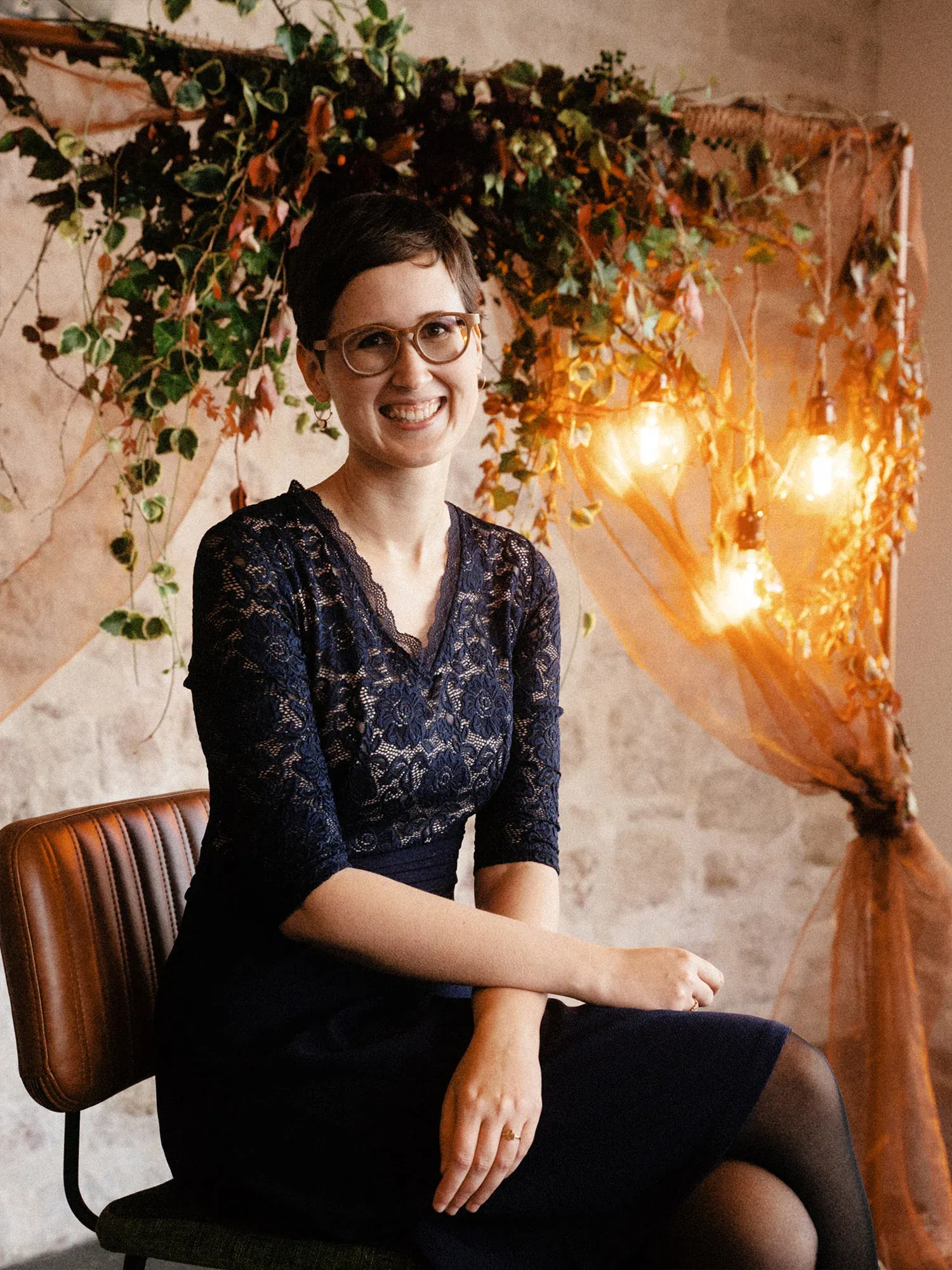 Eine Frau in einem eleganten, dunkelblauen Kleid sitzt lächelnd auf einem Stuhl vor einer herbstlichen Blumendekoration mit warmem Licht.