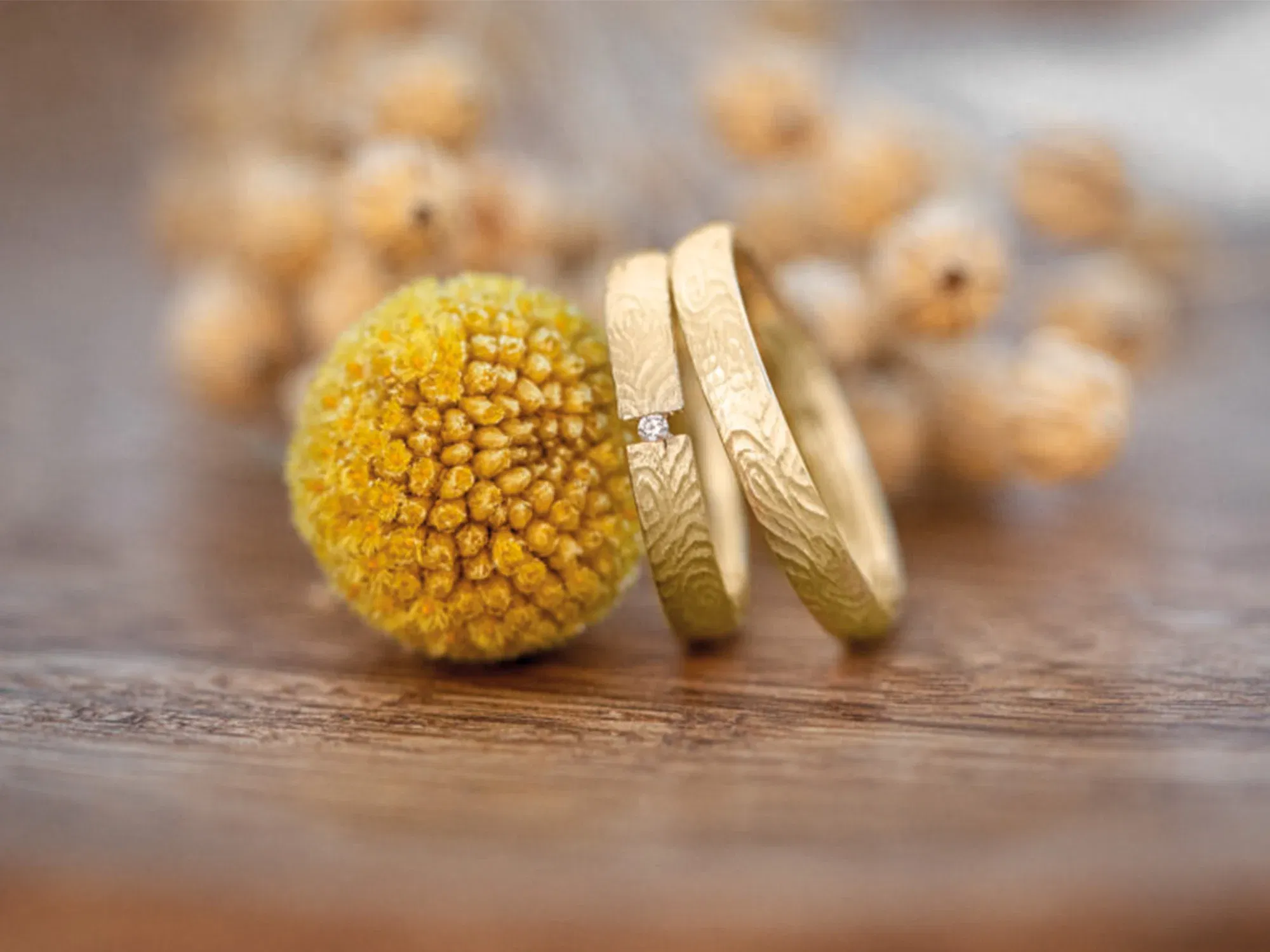 Elegante, goldene Eheringe mit einzigartigem Muster und einem funkelnden Stein, liebevoll arrangiert neben einer dekorativen gelben Blüte.