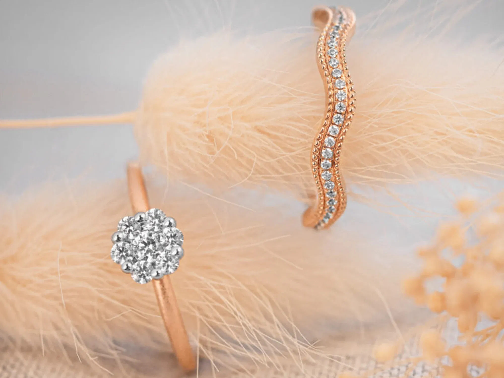Zwei luxuriöse Ringe in Roségold: Ein geschwungener Ring mit einer Reihe kleiner Diamanten und ein schlichter Ring mit einem auffälligen, runden Diamantcluster, dekorativ auf weichem, beigem Material drapiert.