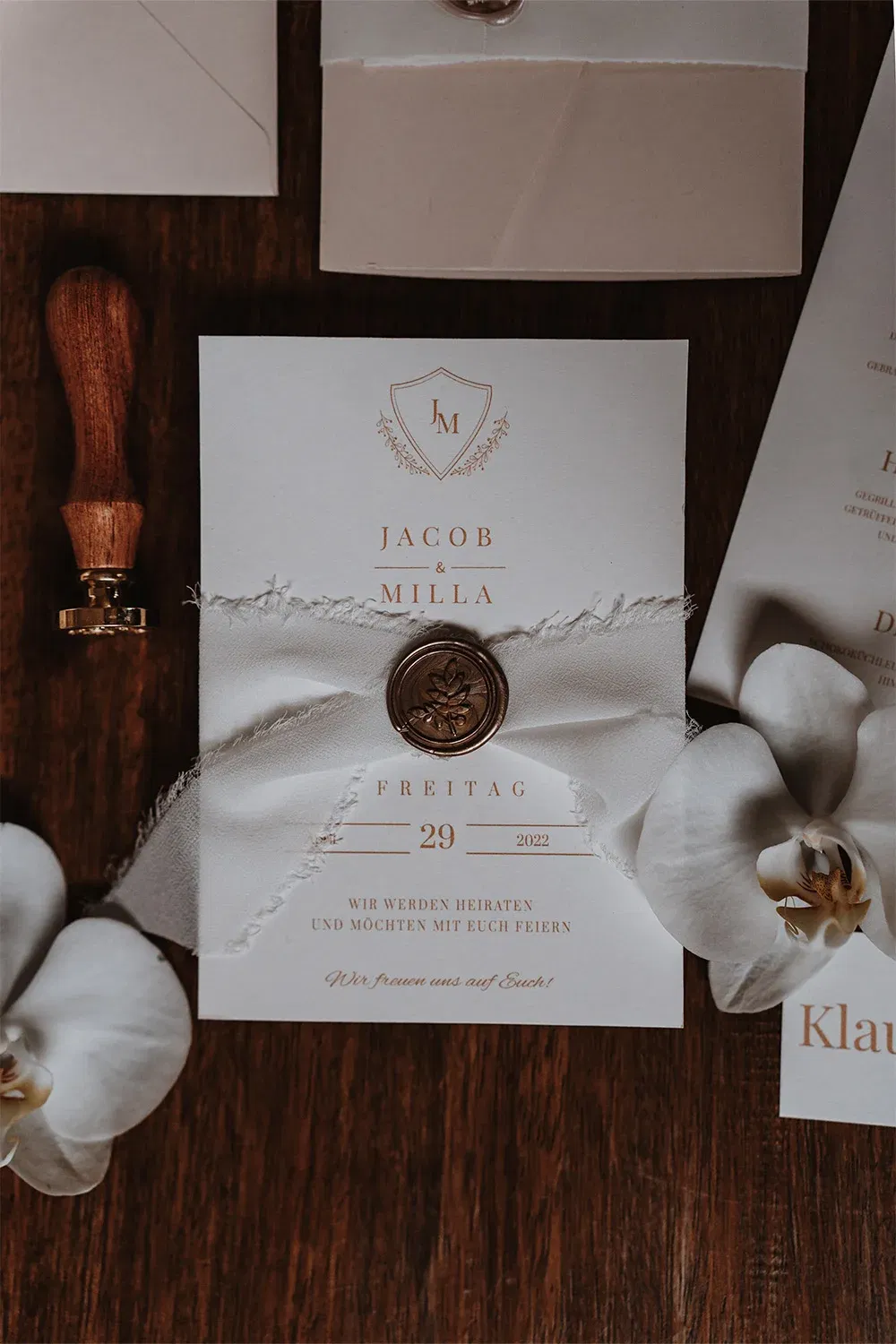 Hochzeitseinladung in edlem Design mit goldener Prägung und einem Wachssiegel. Die Karte ist mit einem weißen Band und Blumen dekoriert.