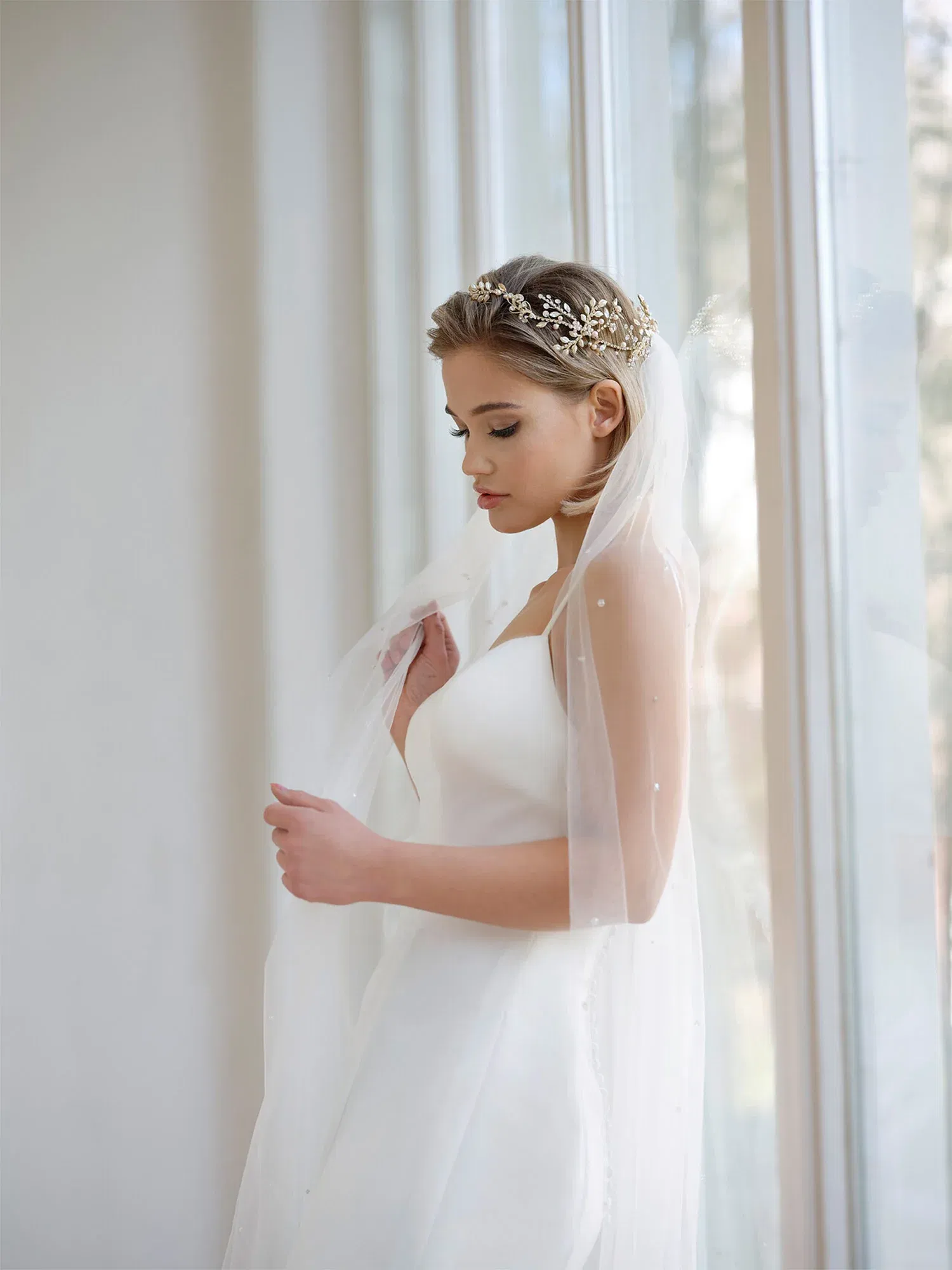 Die Braut in einem eleganten, trägerlosen Kleid mit einem goldenen Tiara-Kranz und einem Schleier, der sanft über ihre Schultern fällt, steht am Fenster.