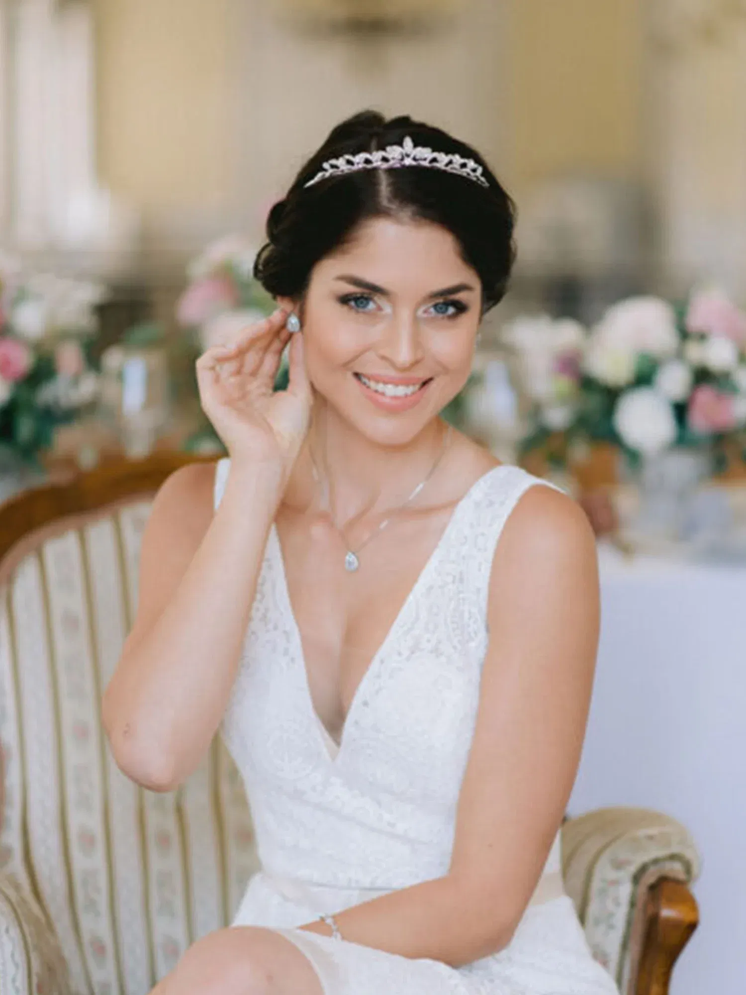 Die Braut sitzt auf einem gepolsterten Stuhl, lächelt, trägt eine elegante Tiara und ein schlichtes, aber stilvolles Spitzenhochzeitskleid.