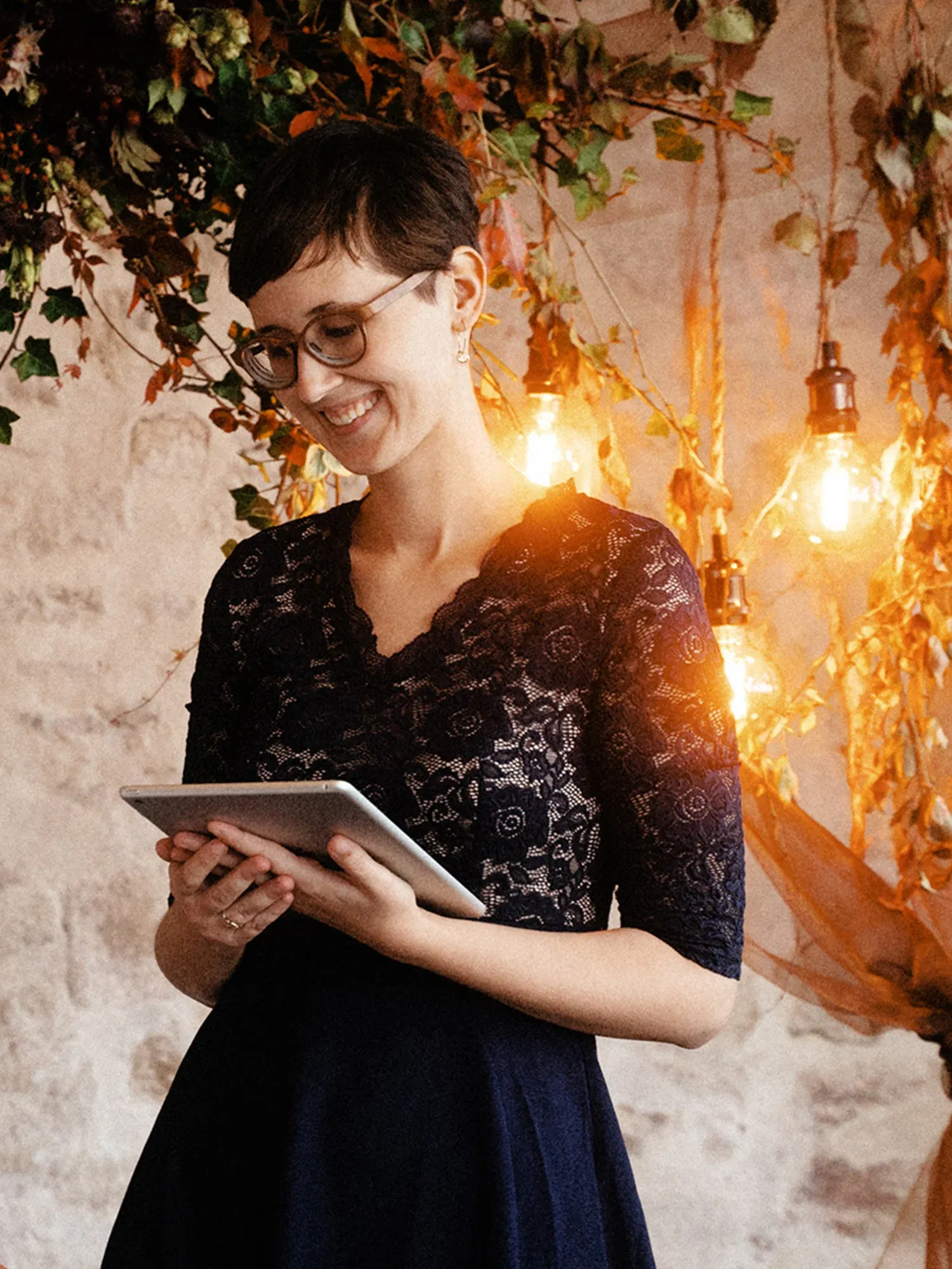 Eine Frau in einem dunkelblauen Spitzenkleid hält ein Tablet in den Händen, umgeben von einer herbstlichen Dekoration und warmem Licht.