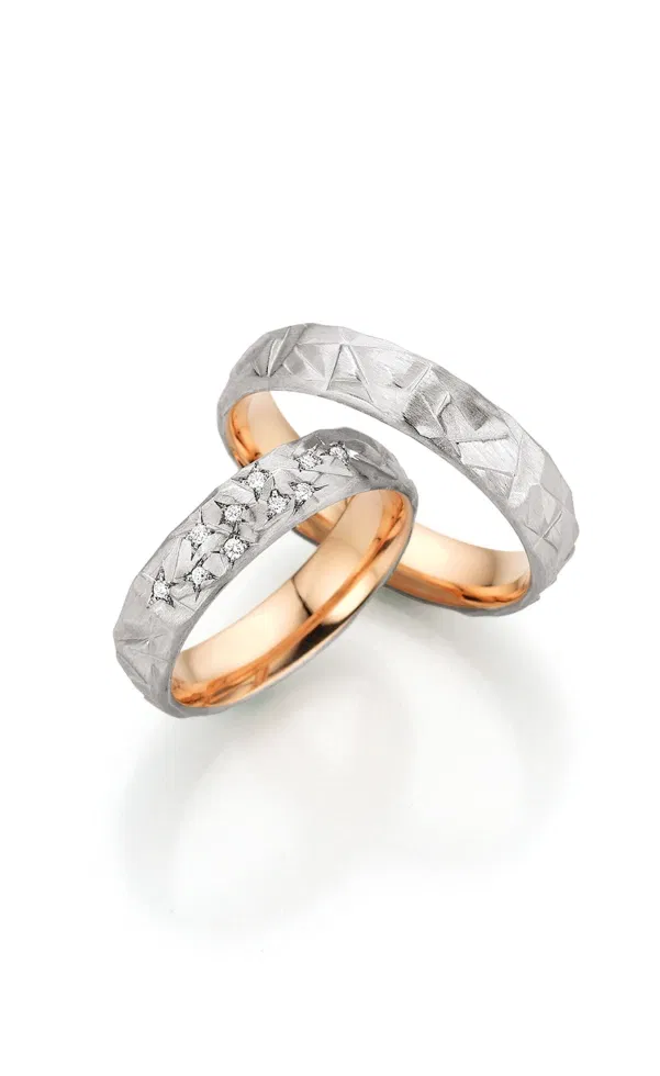 Zwei außergewöhnliche Eheringe aus Weißgold mit strukturierter, eisartig facettierter Oberfläche und warmem Roségold-Innenband, einer davon mit sternförmig eingefassten Diamanten, markant vor hellem Hintergrund inszeniert.