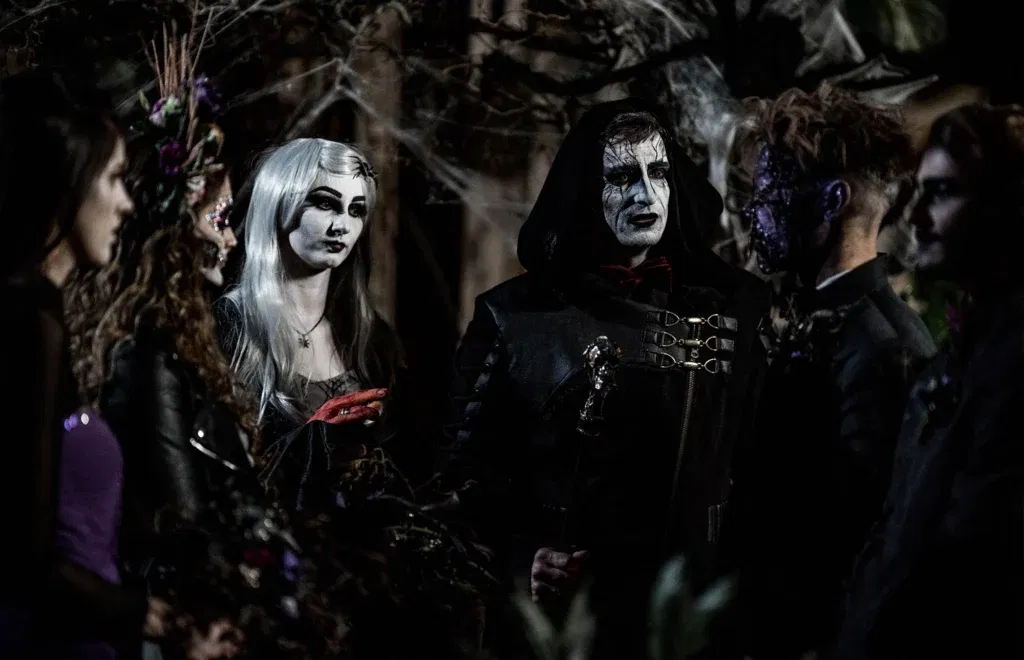 Eine Gruppe von Hochzeitsgästen in dunkler, Gothic-Schminke und Kostümen versammelt sich zu einer unheimlichen Zeremonie, umgeben von Spinnweben und Schatten.