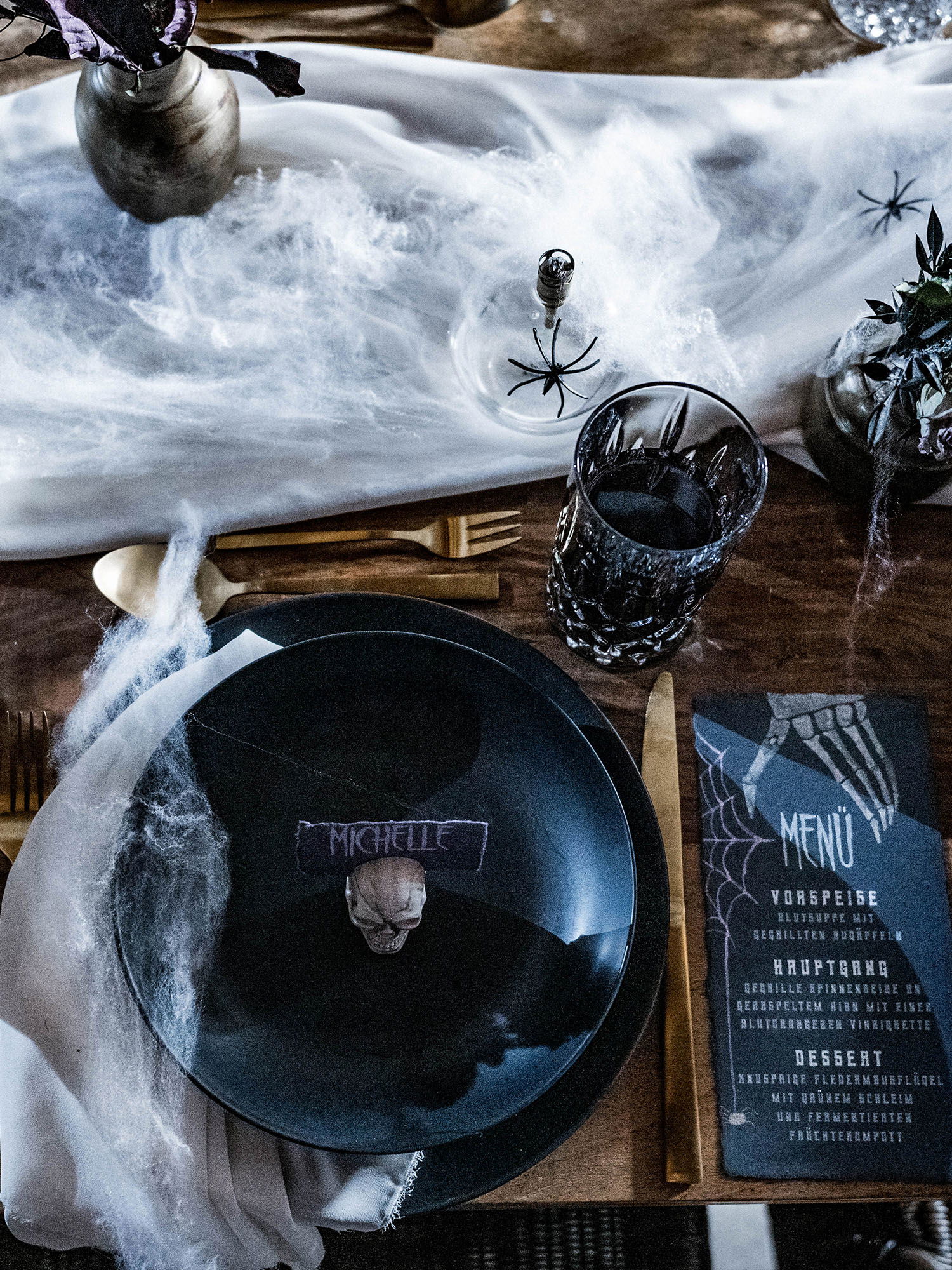 Tischdekoration im Gothic-Stil mit einem schwarzen Teller und Totenkopf, begleitet von einem Menü mit Spinnennetz-Motiven. Spinnweben umgeben das Gedeck.