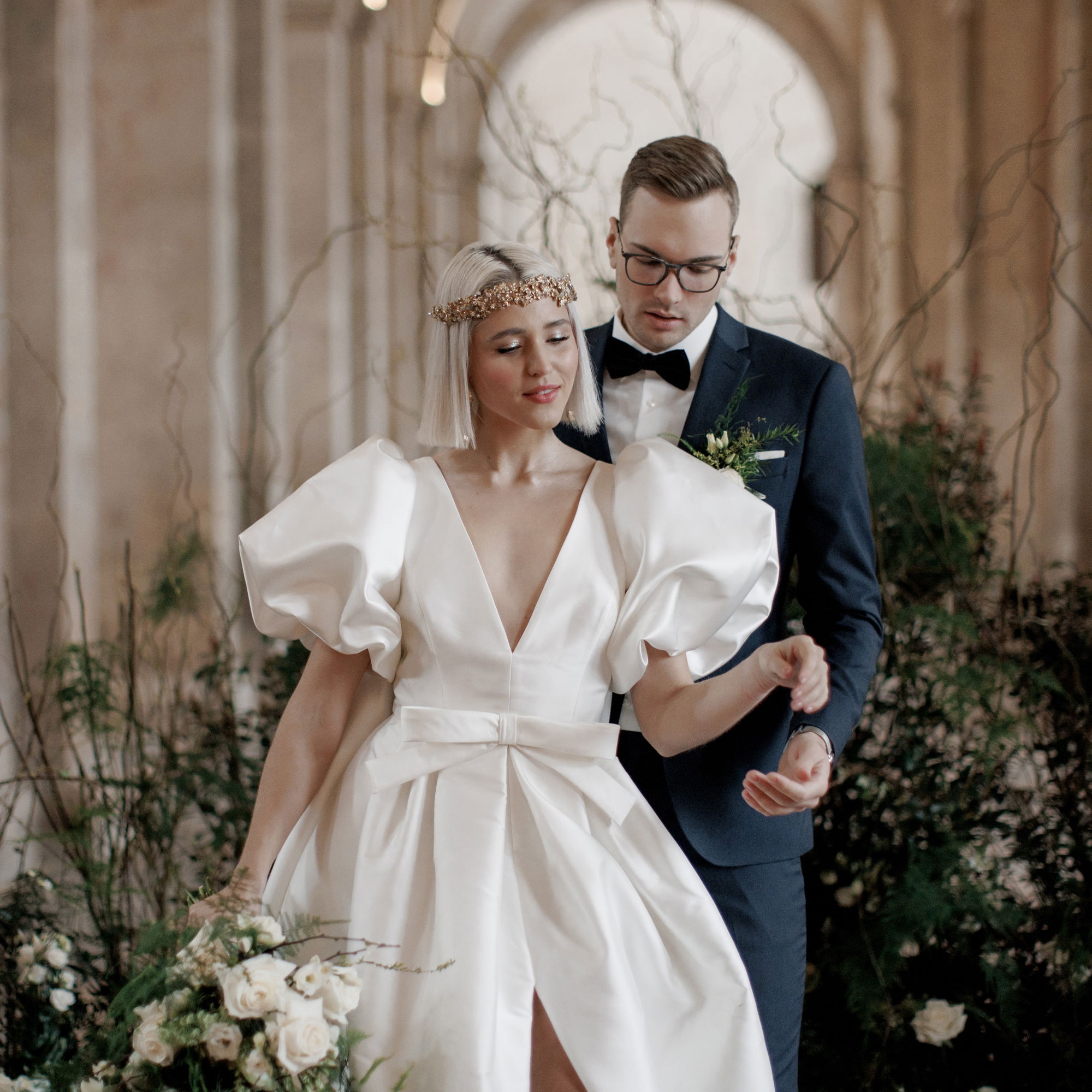 Eine stilvolle Braut in einem weißen Kleid mit voluminösen Ärmeln und ein Bräutigam in einem blauen Anzug posieren für Hochzeitsfotos in einer schicken Location.