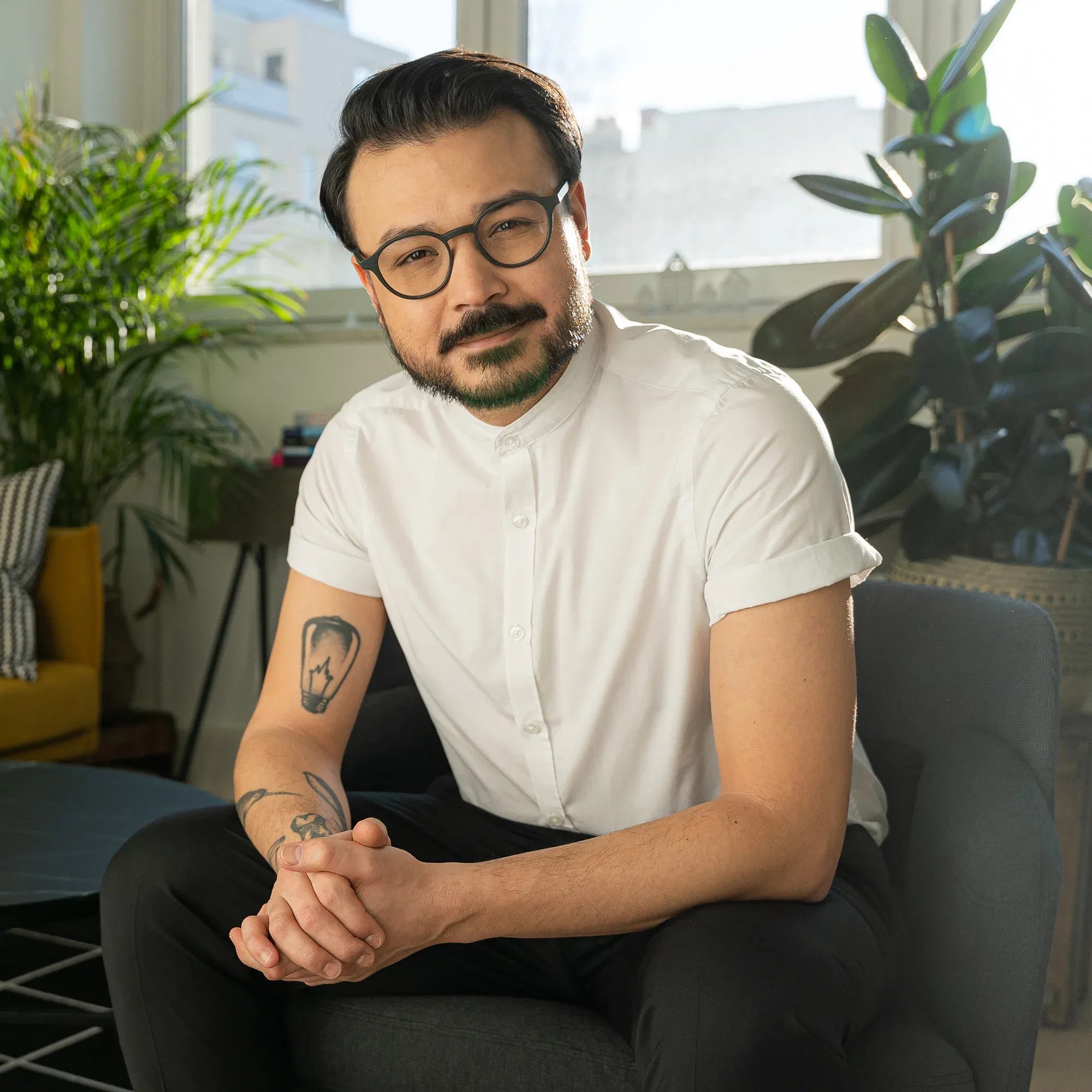 Umut C. Özdemir, lächelnd und in einem weißen Hemd, sitzt in einem gemütlichen Wohnzimmer mit Pflanzen im Hintergrund und blickt in die Kamera.