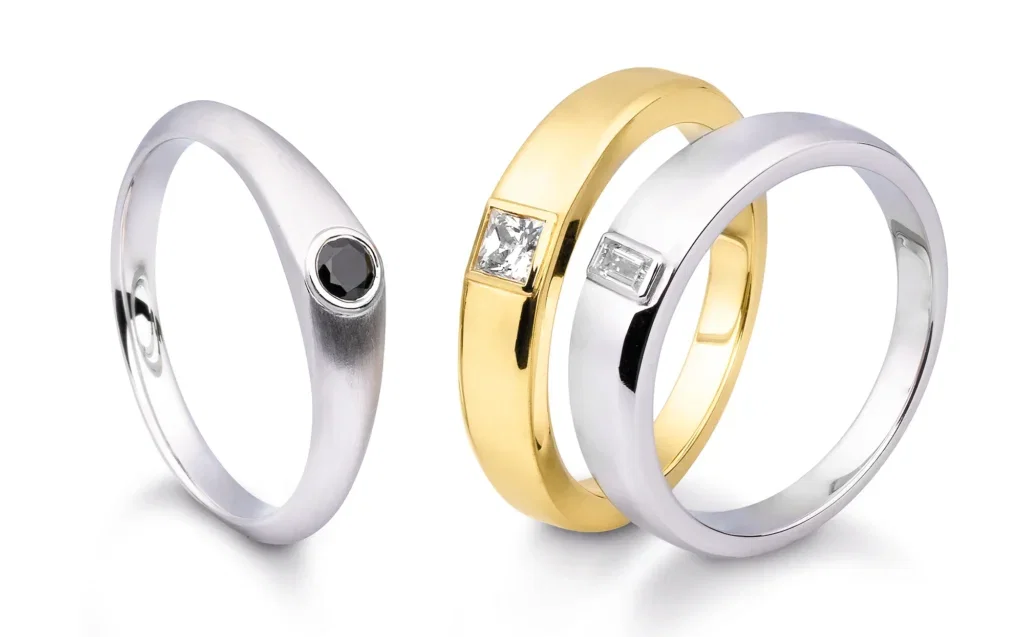 Drei moderne Eheringe, einer in Silber mit schwarzem Stein, und zwei in Gold und Silber mit quadratischen Diamanten als Verzierung.