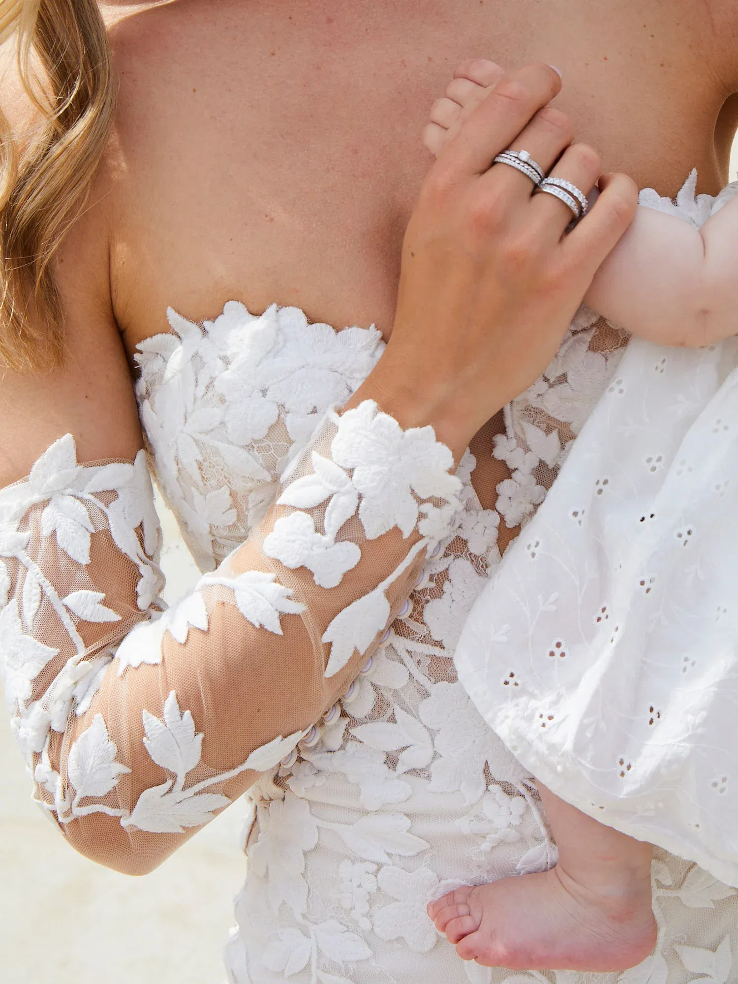 Detailaufnahme einer Braut mit einem Baby. Die Braut trägt ein florales Spitzenkleid, ihr Baby berührt zärtlich ihre Hand, während der Ehering hervorblitzt.