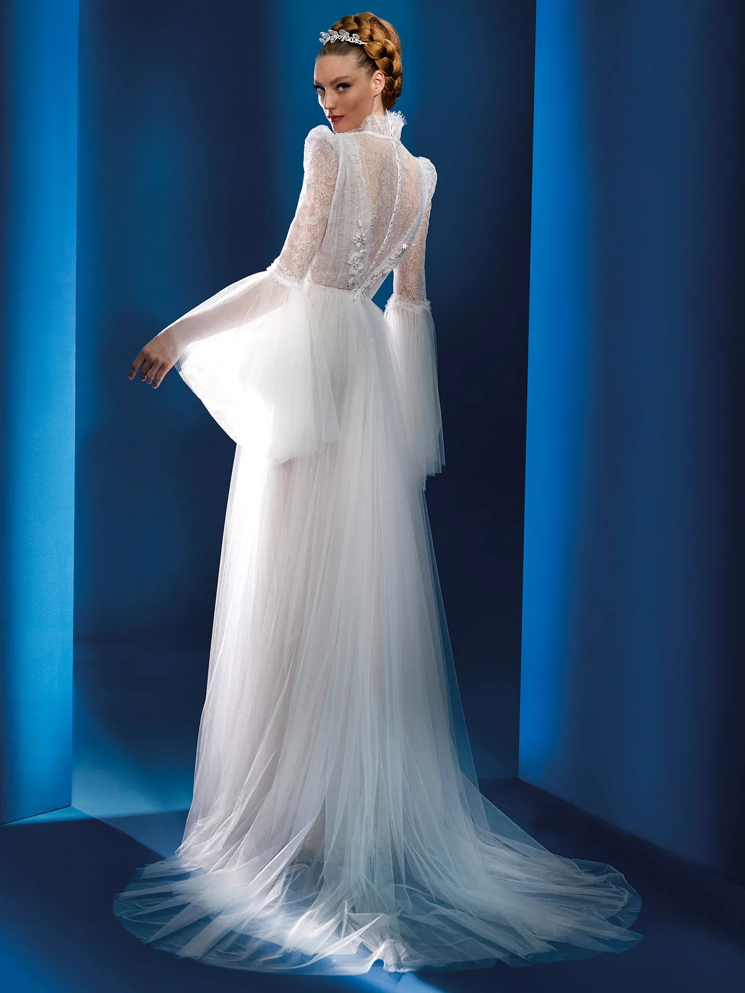 Elegantes Brautkleid mit durchsichtigen Spitzenärmeln und einer langen, fließenden Schleppe aus Tüll. Der hohe Kragen und die dekorativen Knöpfe am Rücken verleihen dem Kleid einen Hauch von klassischer Eleganz, während die weiten Ärmel moderne Romantik ausstrahlen.