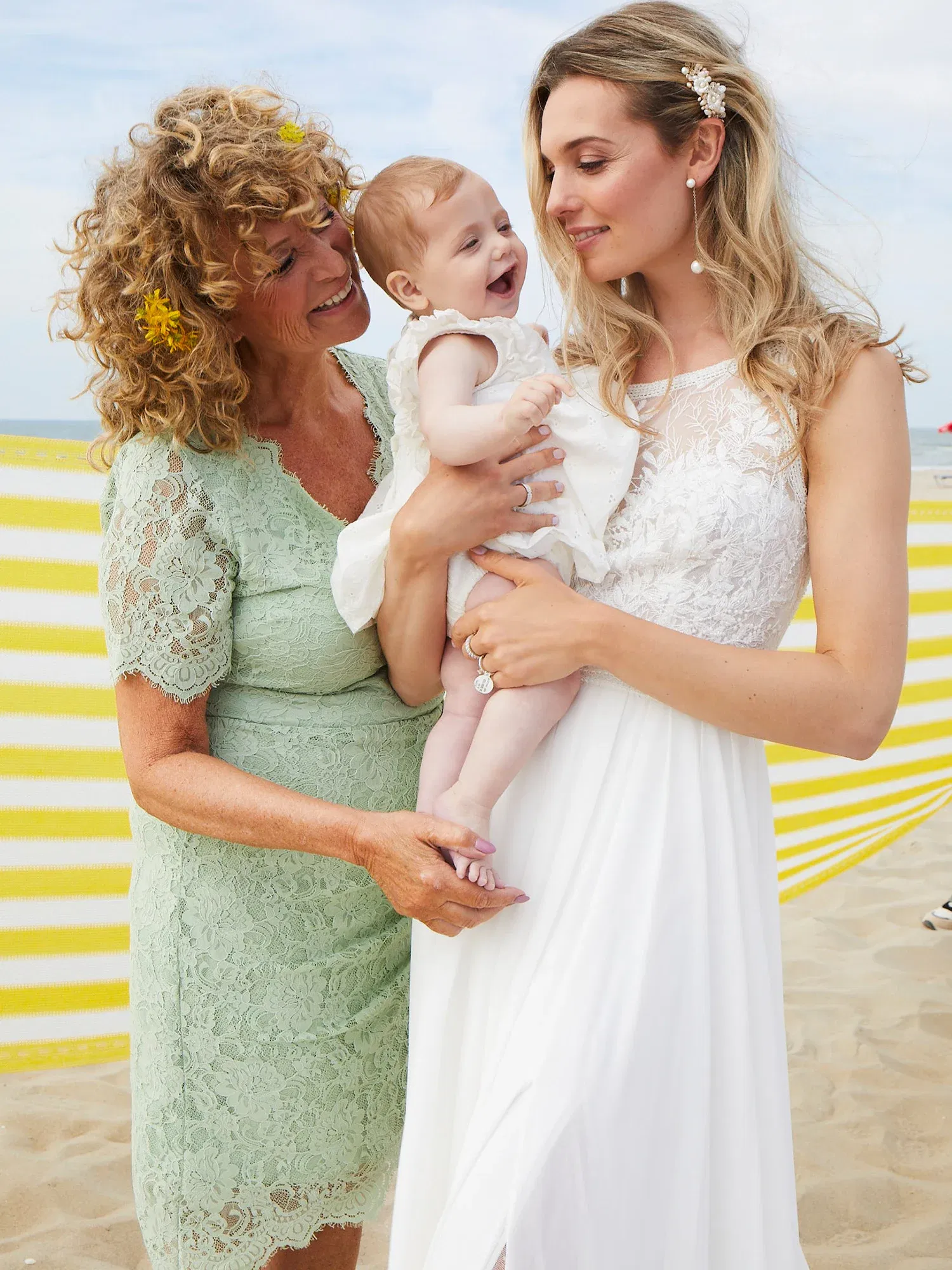 Ein fröhliches Strandfoto mit Großmutter, Braut und Baby. Die Braut trägt ein weißes Kleid, während die Großmutter in einem mintfarbenen Spitzenkleid das Baby liebevoll berührt.