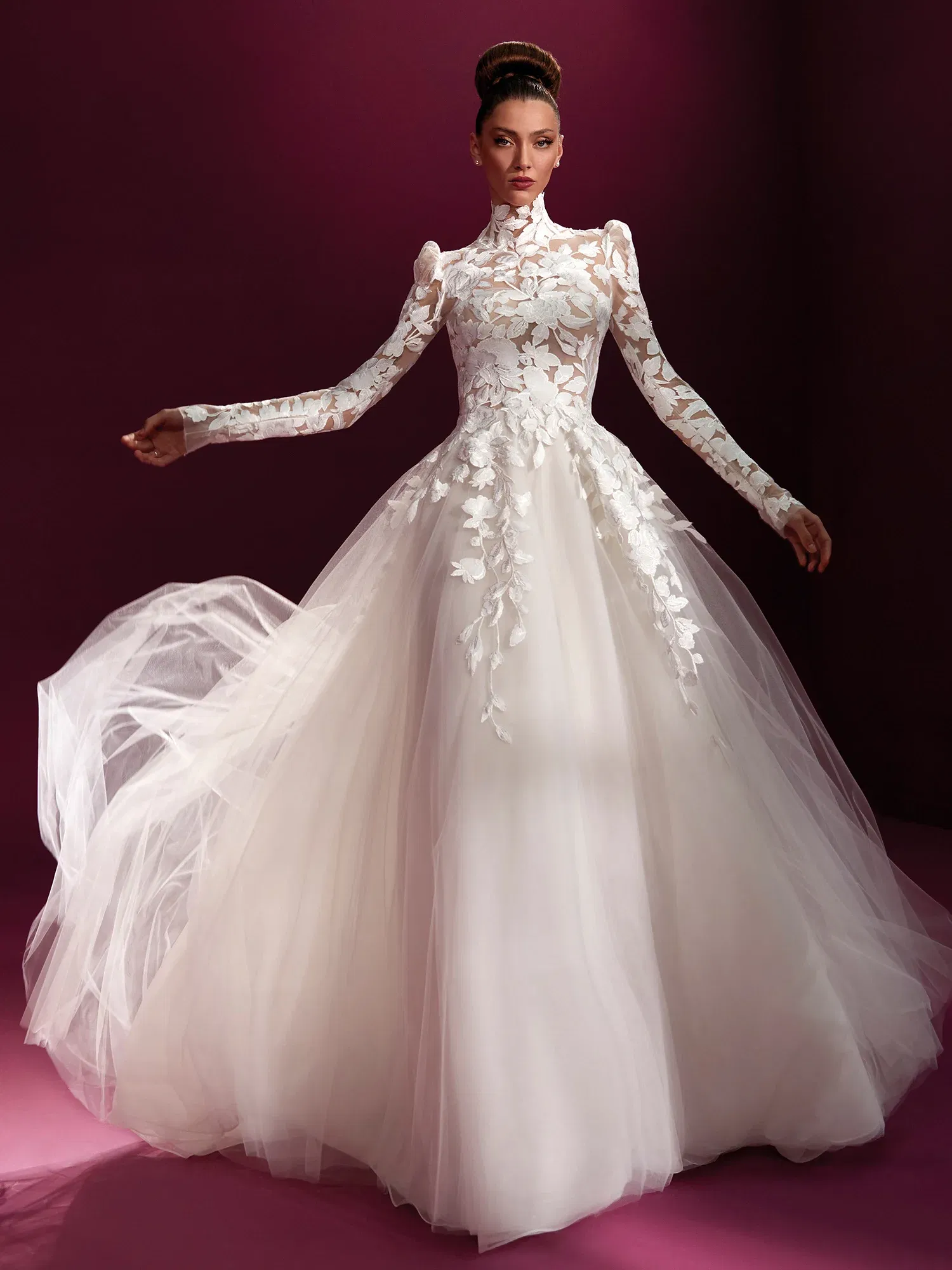 Ein prunkvolles Brautkleid mit transparenten Spitzenärmeln und hohem Kragen, das eine Mischung aus moderner Eleganz und klassischer Romantik ausstrahlt. Der weite Tüllrock unterstreicht die majestätische Ausstrahlung des Outfits.