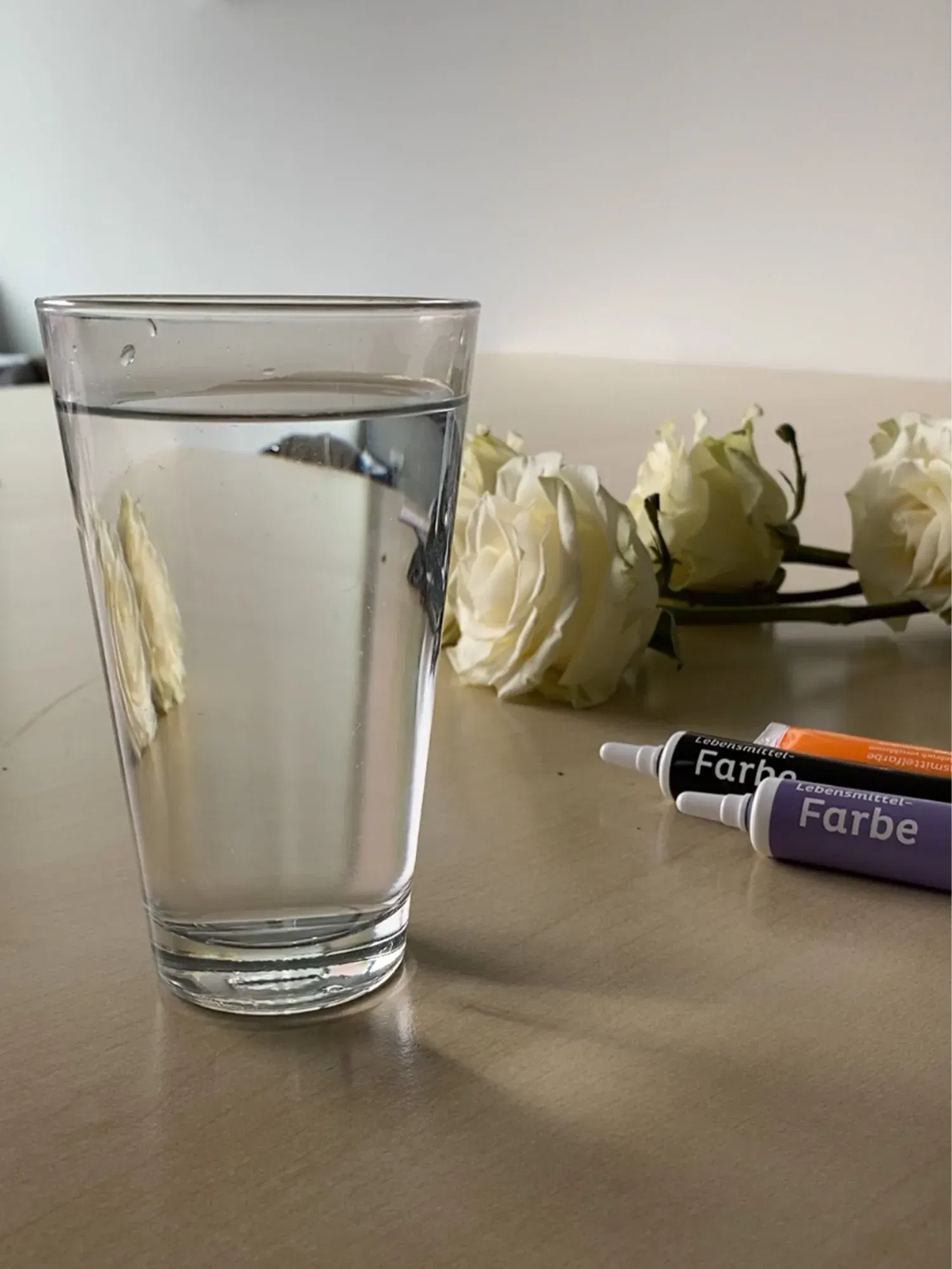 Ein Glas klares Wasser auf einem Holztisch, umgeben von weißen Rosen. Lebensmittelfarben-Tuben liegen daneben und deuten auf ein kreatives Experiment hin.
