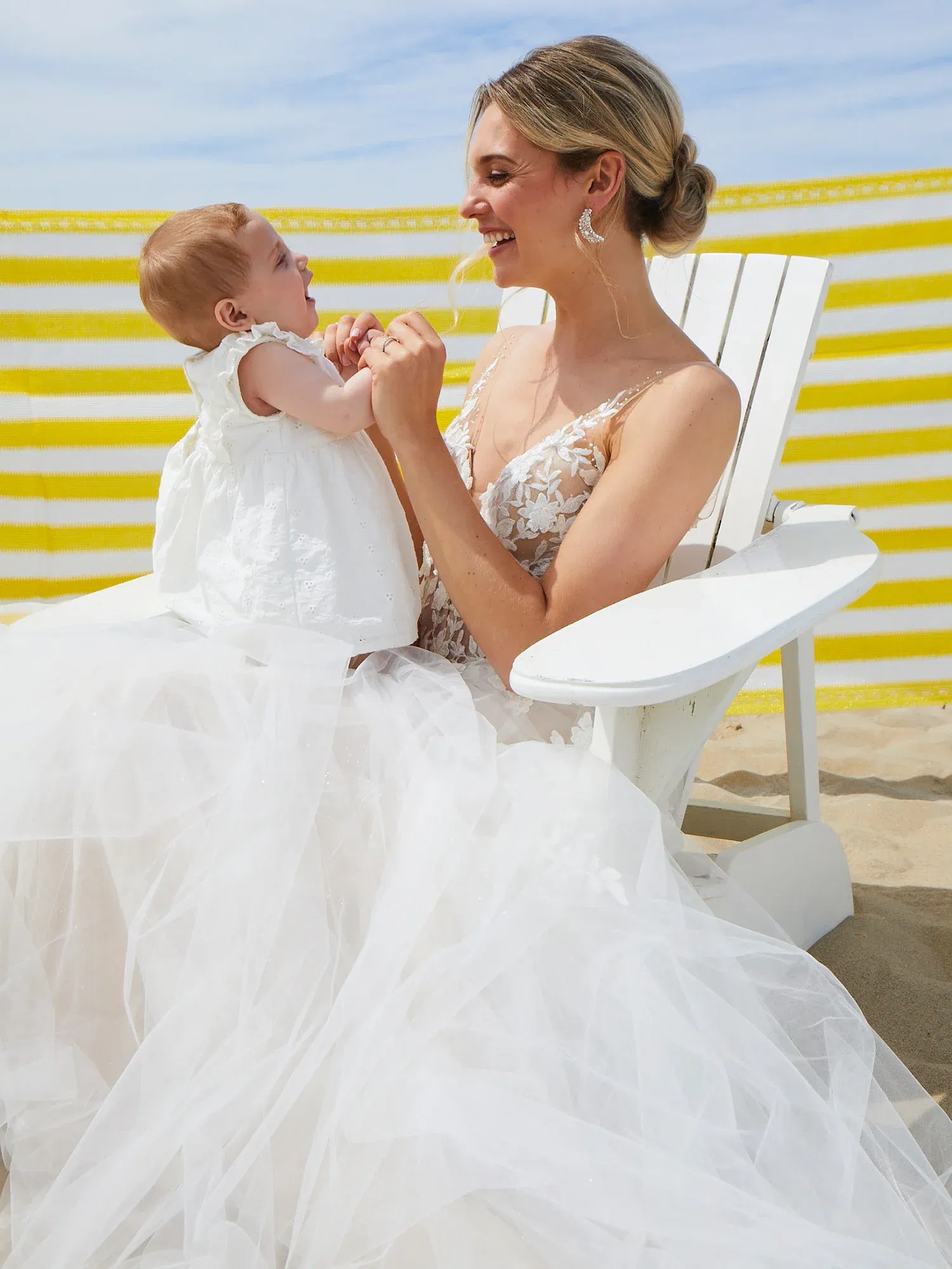 Eine Braut in einem großen, fließenden Hochzeitskleid sitzt auf einem weißen Stuhl am Strand und spielt fröhlich mit ihrem Baby auf dem Schoß.