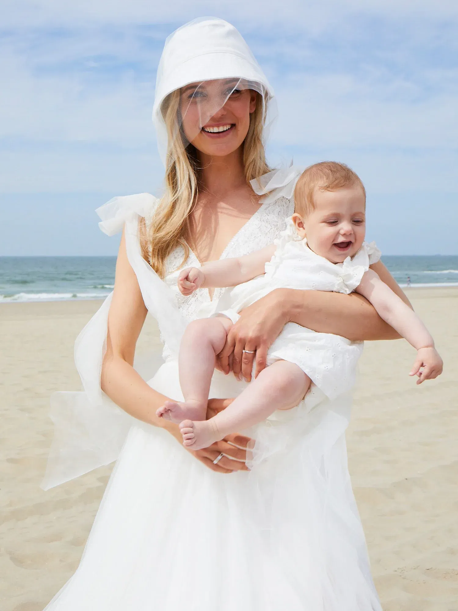 Strahlende Braut mit weißem Kleid und passendem Hut hält ihr Baby. Das Baby lacht, während sie gemeinsam den Strand genießen.