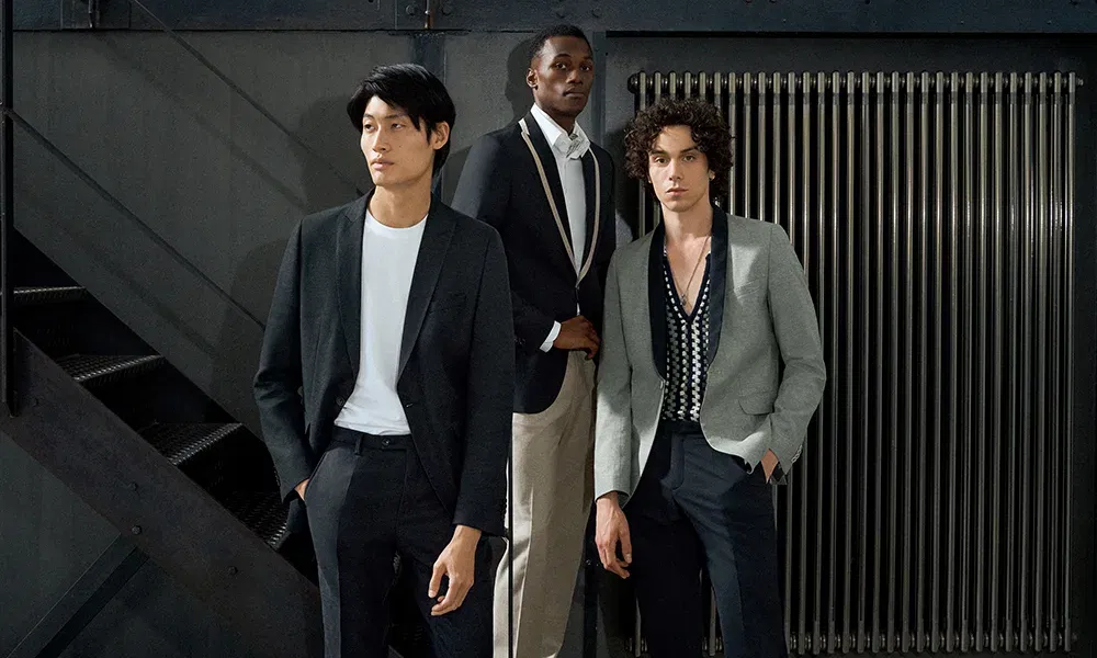 Eine Gruppe von drei Männern posiert vor einer metallischen, industriellen Kulisse. Der Mann links trägt einen minimalistischen schwarzen Anzug mit einem weißen T-Shirt, während der Mann in der Mitte in einem eleganten dunkelblauen Blazer und beigen Hosen gekleidet ist. Der Mann rechts hebt sich durch seinen grauen Blazer und das gemusterte Hemd hervor. Ihre Outfits vermitteln einen modernen, zeitlosen Stil.