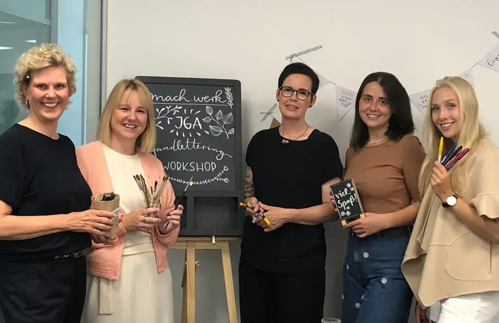 Eine Gruppe von fünf Frauen posiert fröhlich für ein Foto während eines Handlettering-Workshops. Sie halten Werkzeuge zum Schreiben und Gestalten in der Hand, während hinter ihnen eine Tafel mit kreativer Schriftgestaltung steht.