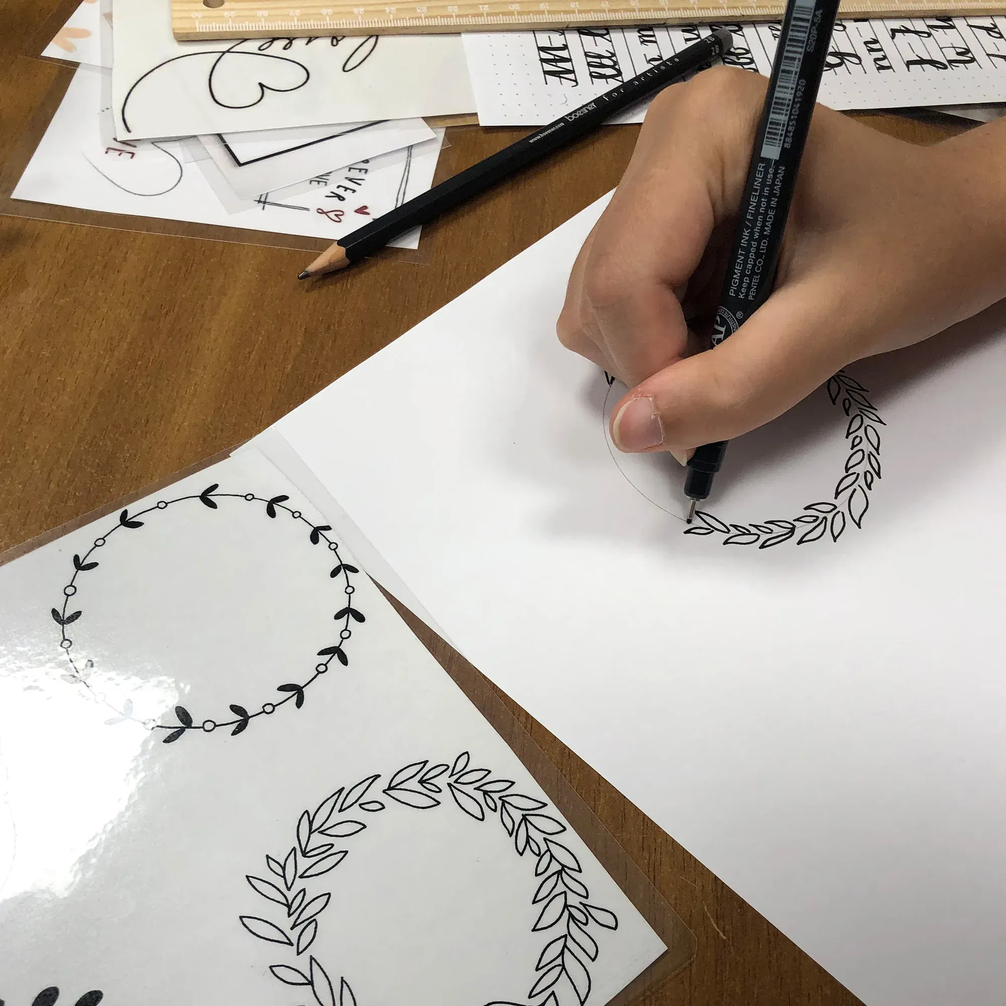 Eine Person gestaltet einen filigranen Blätterkranz mit einem schwarzen Stift auf einem Blatt Papier. Auf dem Tisch befinden sich mehrere Handlettering-Vorlagen mit ähnlichen floralen Motiven.