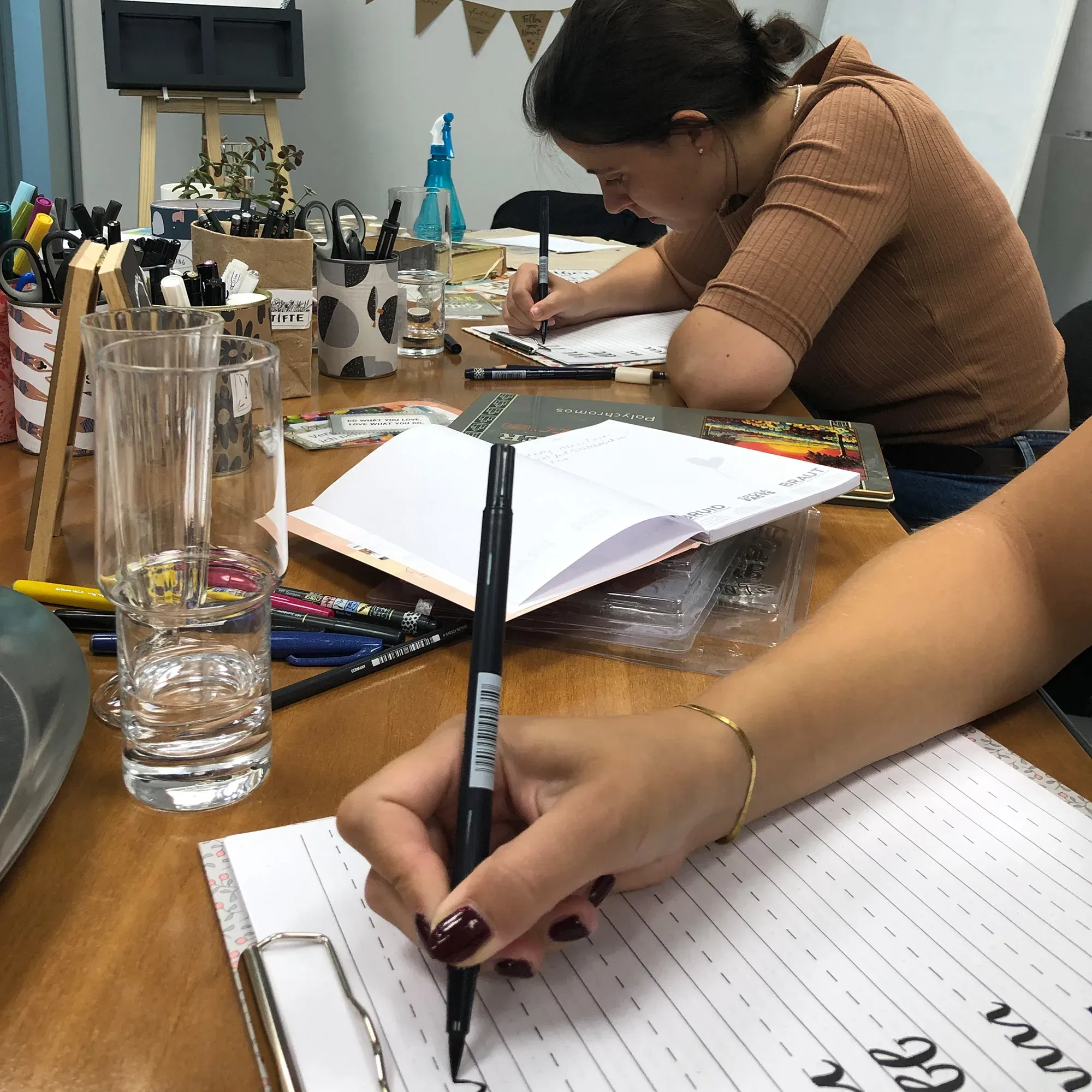 Eine Teilnehmerin eines Handlettering-Workshops konzentriert sich auf ihre Übungen, während sie mit einem schwarzen Pinselstift Buchstaben auf ein Übungsblatt schreibt. Der Tisch ist mit verschiedenen Stiften und Utensilien bedeckt.