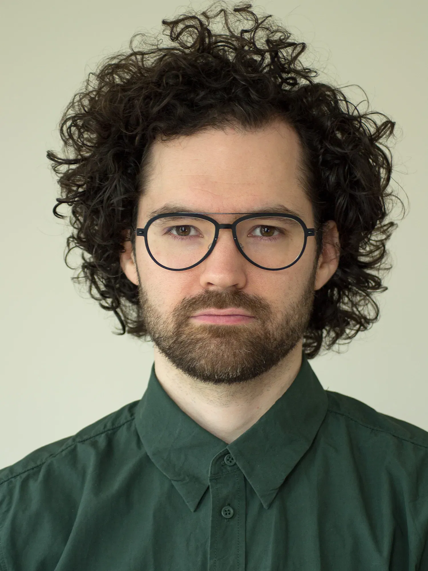 Nahaufnahme von Lukas Grewenig, der mit Brille, lockigem Haar und einem dunklen Hemd in die Kamera schaut, vor neutralem Hintergrund.