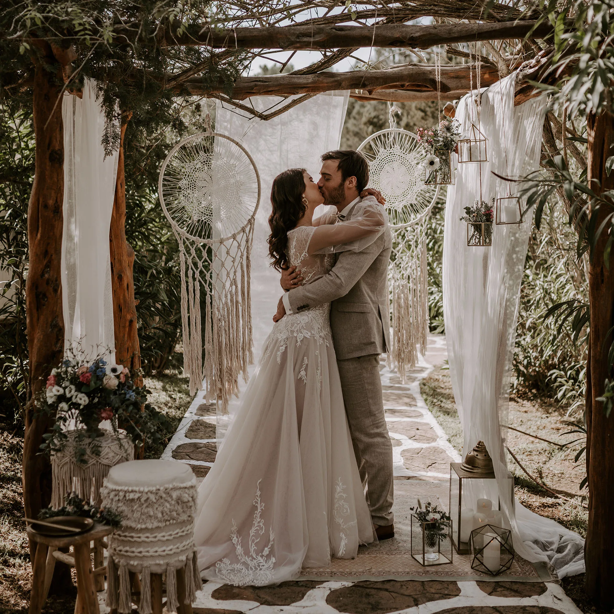 Das Brautpaar umarmt sich unter einem rustikalen Bogen mit Traumfängern und floralen Dekorationen während ihrer Boho-Hochzeit im Freien.