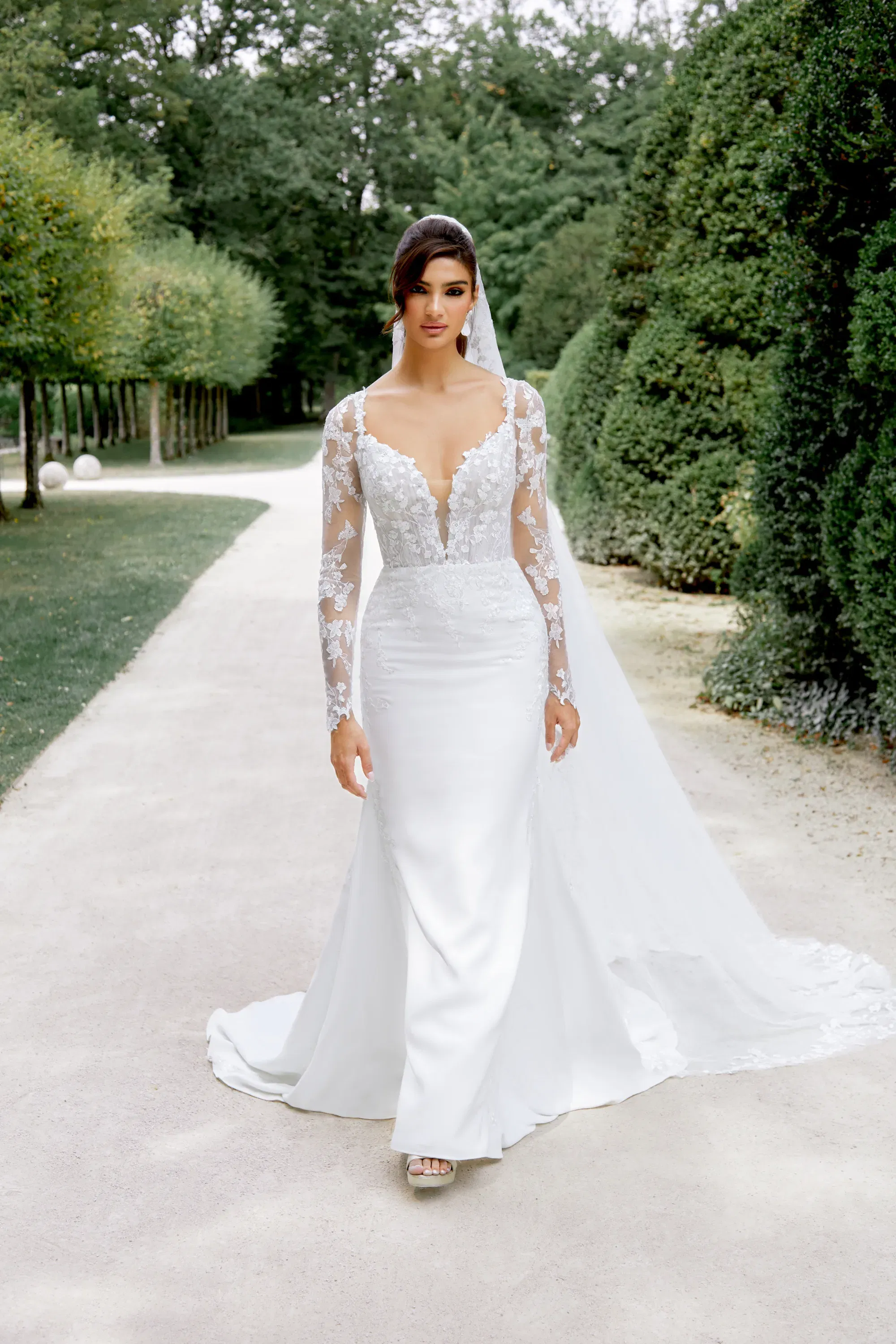Fit-and-Flare-Brautkleid mit tiefem V-Ausschnitt und langen Spitzenärmeln, kombiniert mit einem langen Schleier – präsentiert auf einem Alleeweg.