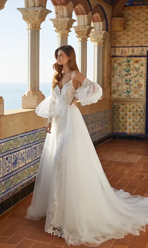 In einem traumhaften Kleid mit luftigen, durchsichtigen Ärmeln und filigranen Spitzenverzierungen posiert die Braut auf einem Balkon mit Blick aufs Meer. Die dekorativen Kacheln und Säulen des historischen Gebäudes unterstreichen den romantischen Charme des Outfits.