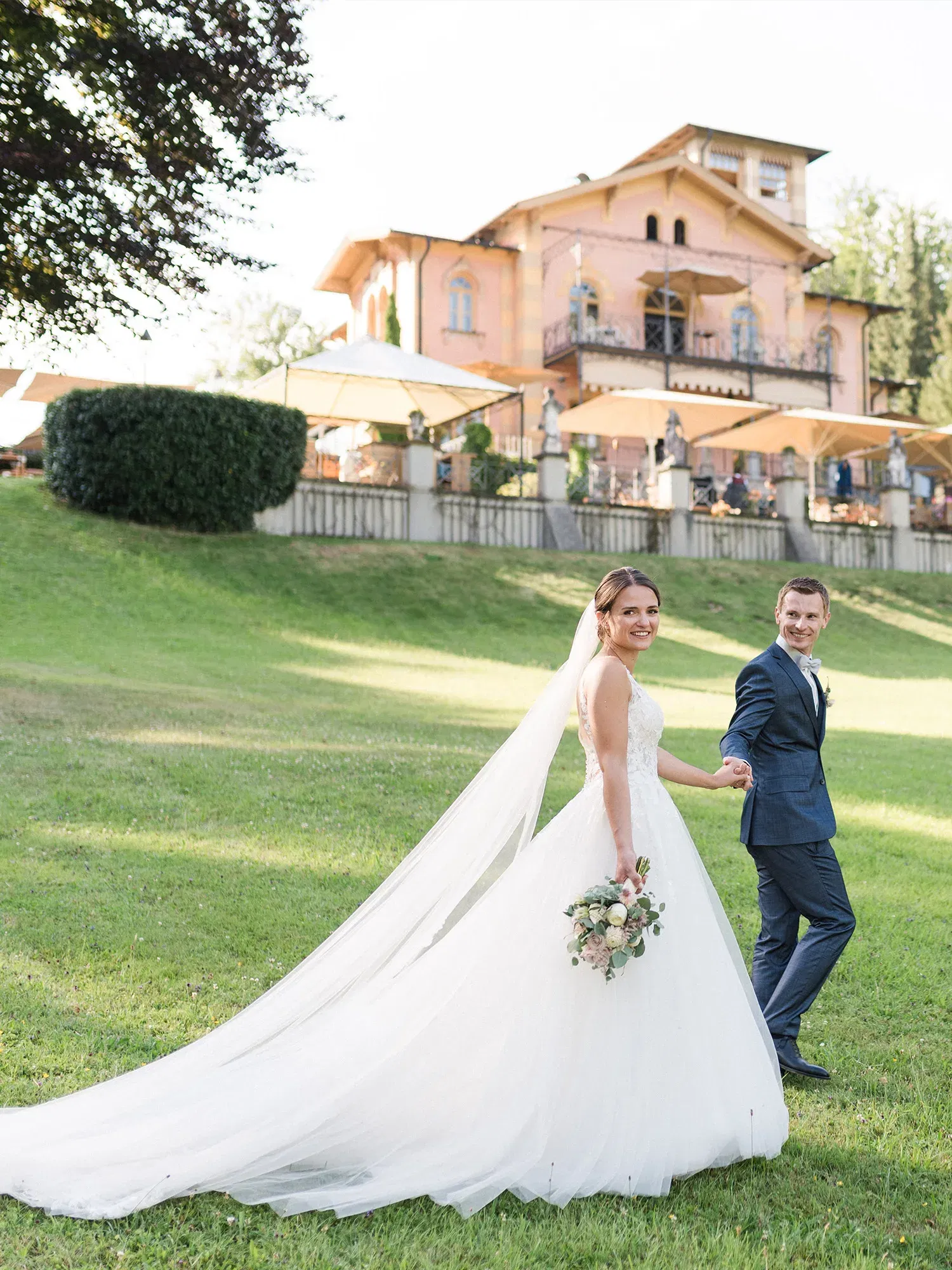 Das Brautpaar genießt einen Moment in der Natur vor einem rosafarbenen Herrenhaus. Die Braut in einem bodenlangen, weißen Kleid mit Schleppe lächelt über die Schulter, während ihr Ehemann ihre Hand hält.