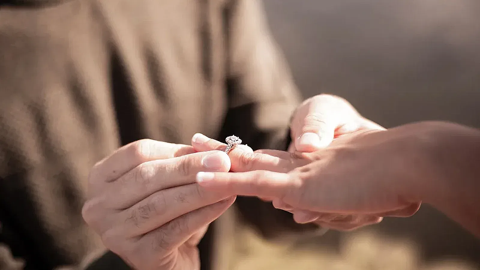 Detailansicht einer Verlobung, bei der ein funkelnder Diamantring an den Finger einer Person gesteckt wird.
