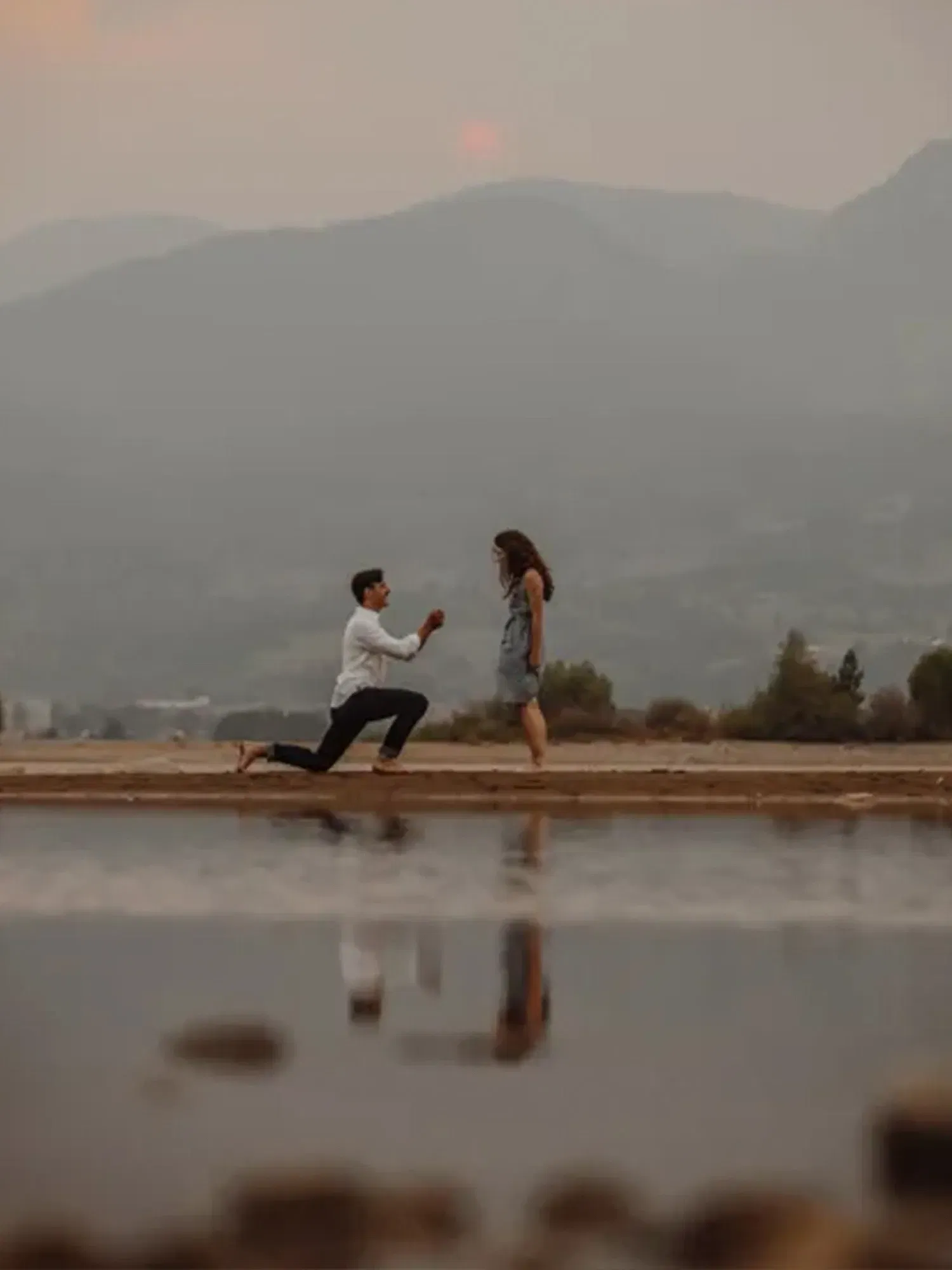 Ein Heiratsantrag im Freien bei Sonnenuntergang: Der Mann kniet nieder und zeigt seiner Partnerin den Verlobungsring.