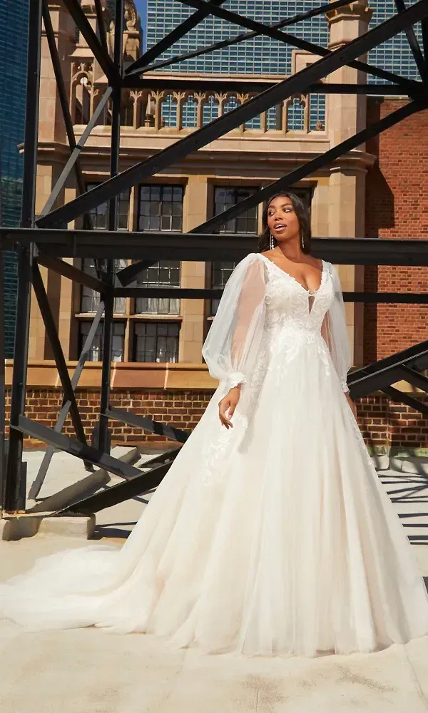 Die Braut in einem fließenden, mit Spitze verzierten Kleid mit Puffärmeln posiert vor einer modernen Kulisse, die von Gebäuden aus Glas und Stahl dominiert wird. Ihr luxuriöses Kleid hebt sich vom industriellen Hintergrund ab.