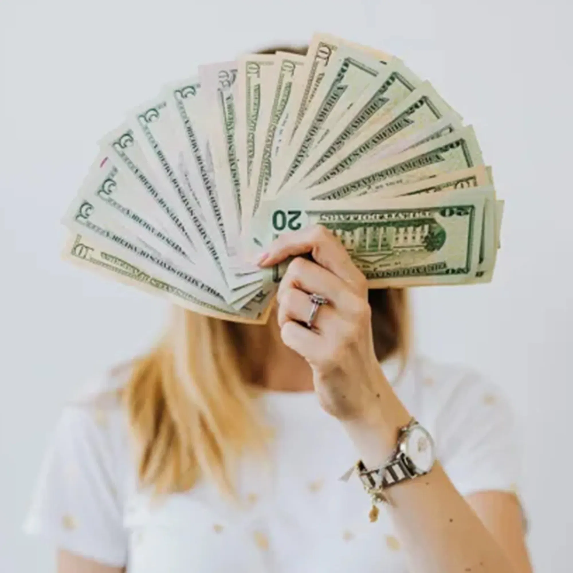 Eine Frau verdeckt ihr Gesicht mit einem Fächer aus Dollar-Scheinen, wobei ihr Arm und die Uhr sichtbar bleiben. Sie trägt ein helles, gemustertes T-Shirt.