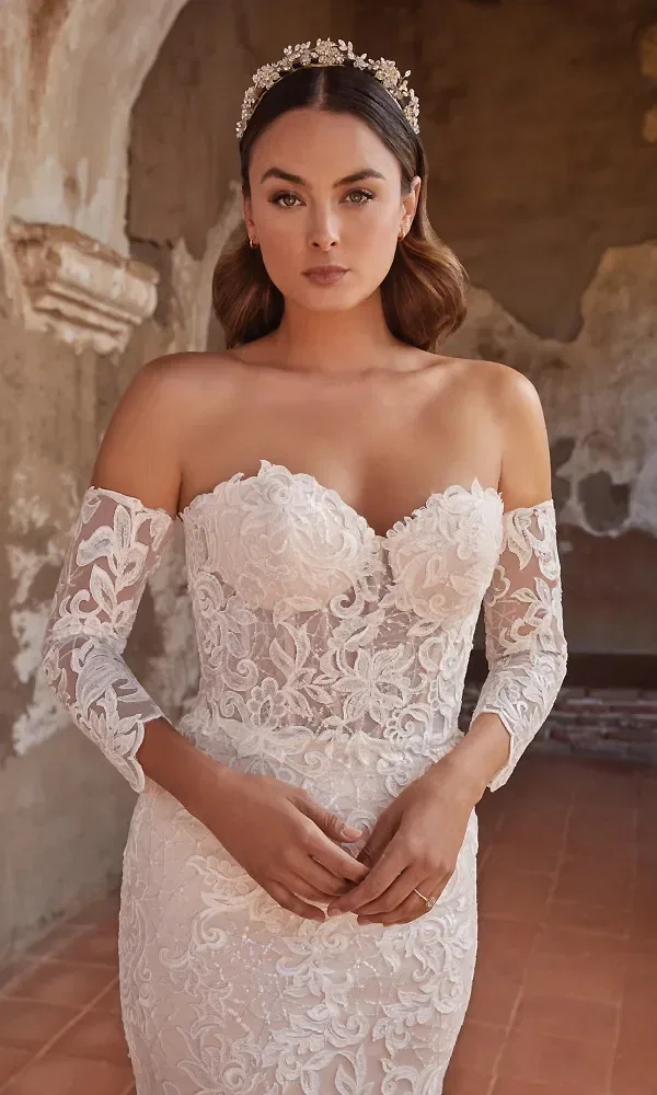 Die Braut trägt ein aufwendig verziertes, trägerloses Kleid mit floralen Spitzenmustern und halbtransparenten Ärmeln. Ihr Haar wird von einem funkelnden Diadem gekrönt, während sie in einer rustikalen Umgebung mit warmem, natürlichem Licht posiert.