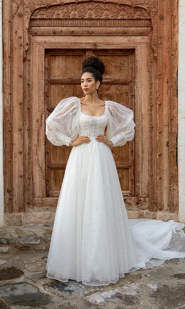 Das Bild zeigt eine Braut in einem voluminösen Brautkleid mit ausgeprägten Puffärmeln und einem Spitzenoberteil. Sie posiert vor einer kunstvoll verzierten Holztür, die dem Gesamtbild einen rustikalen und zugleich traditionellen Charme verleiht.